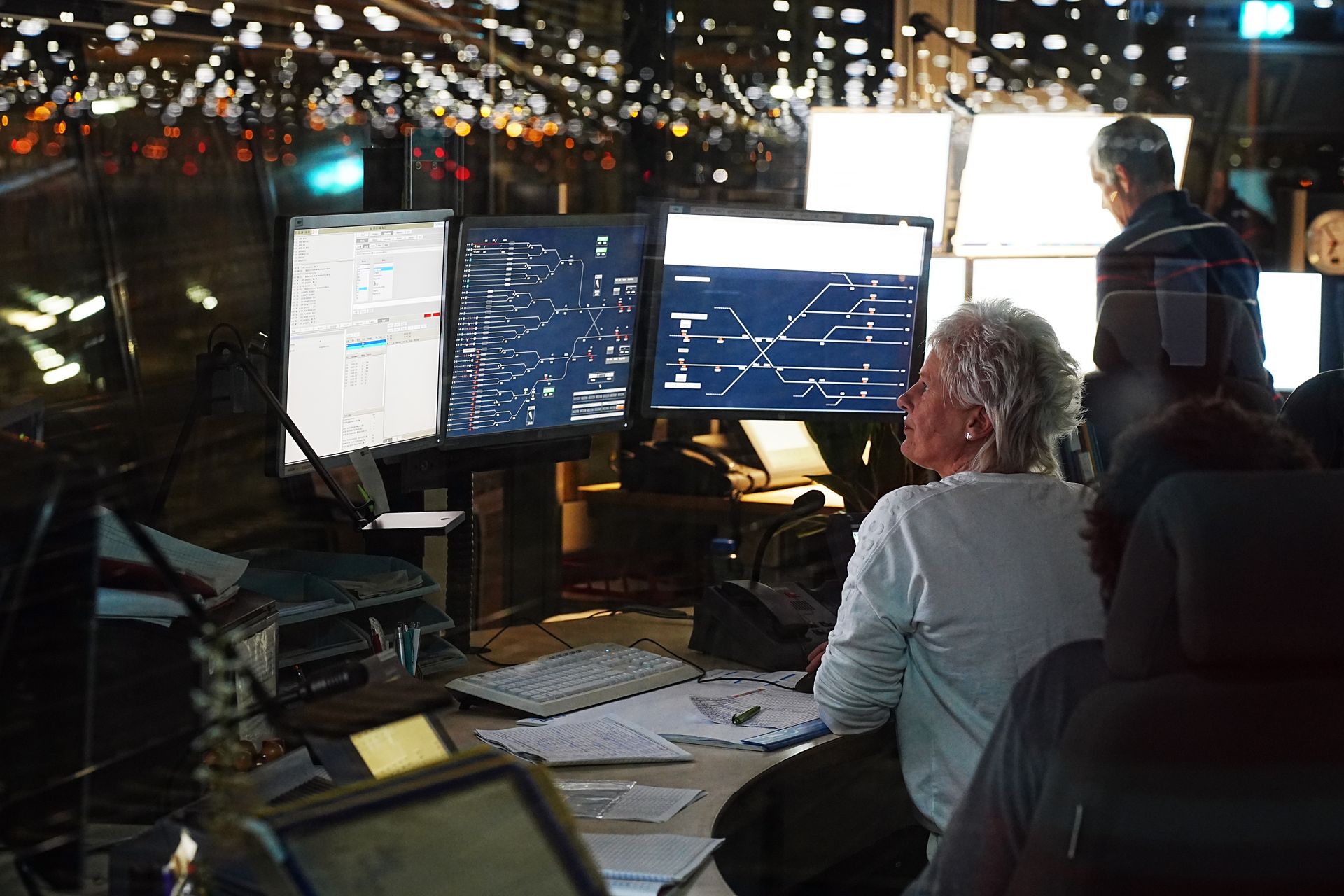 Operators in a control room monitor computer screens displaying complex data visuals and network diagrams, reflecting a busy workspace.