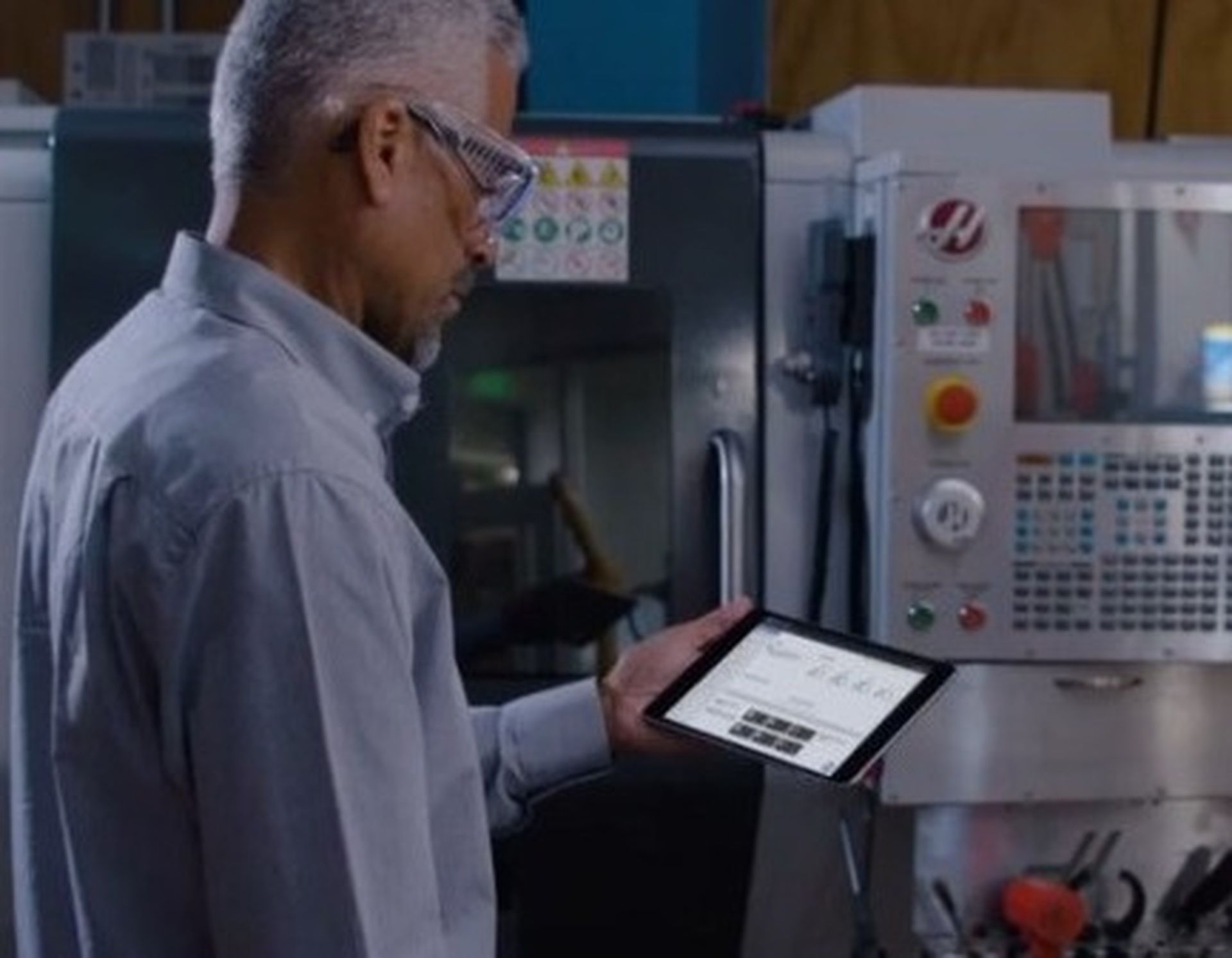 Man in glasses using a tablet near industrial machinery, focusing on the screen with digital controls visible on a CNC machine in the background.