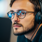Összpontosító fiatal férfi szemüvegben és fejhallgatóban modern irodában dolgozik