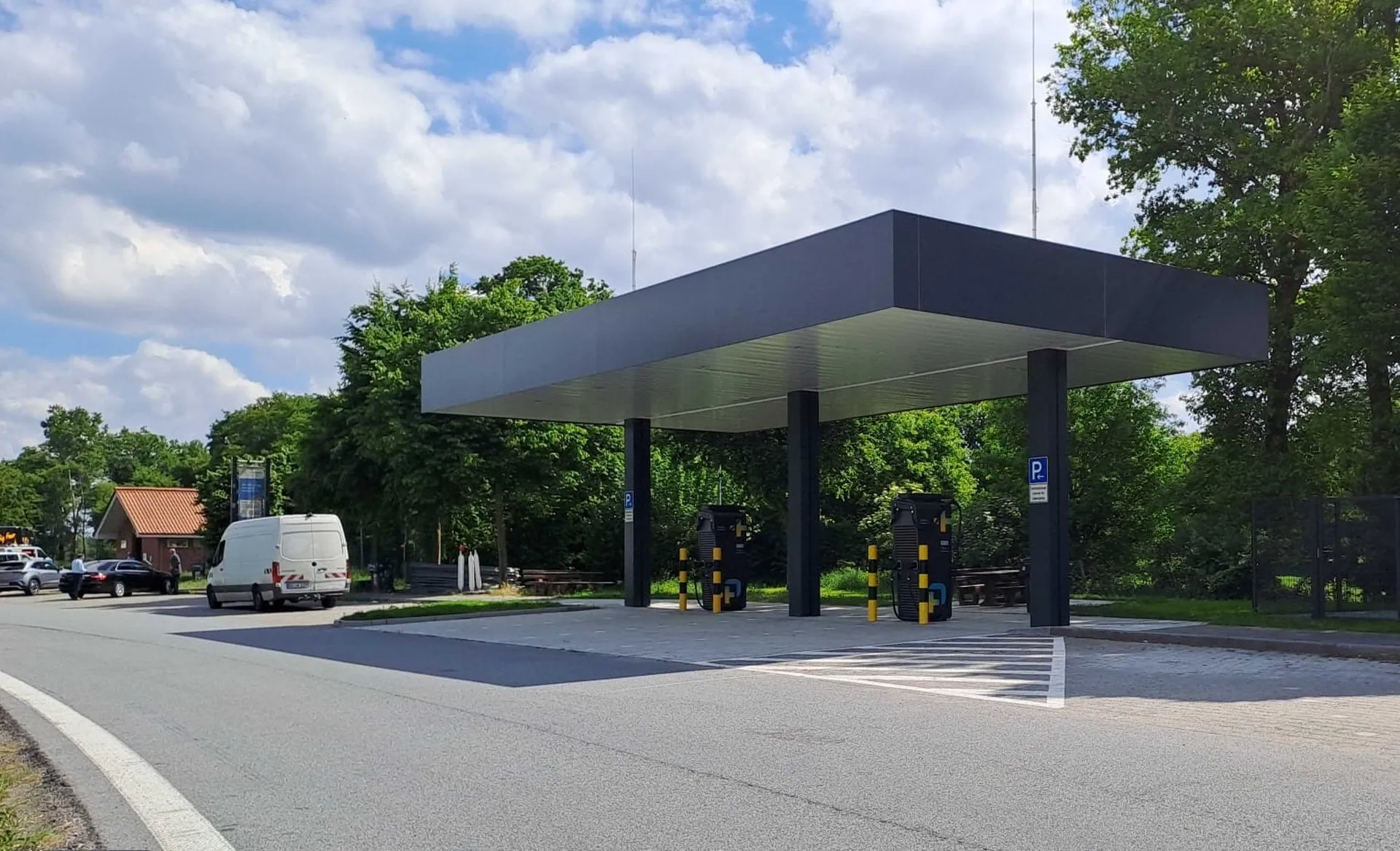 Ein überdachter Ladepark auf einem Autobahn-Parkplatz. Zwei Ladesäulen des Deutschlandnetzes stehen unter einem dunklen Dach. Im Hintergrund sind grüne Bäume und ein bewölkter Himmel zu sehen.