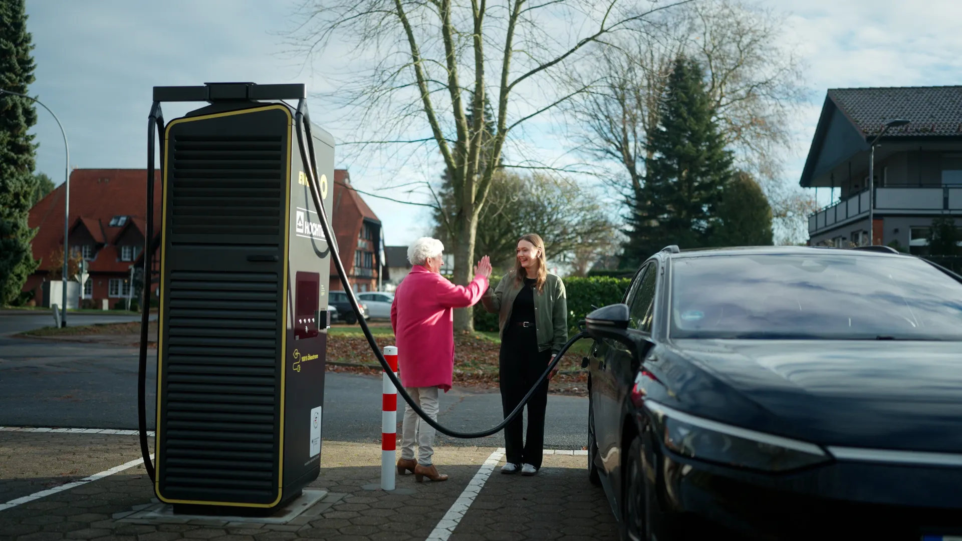 Eine ältere Frau im pinken Mantel und eine jüngere Frau in olivgrüner Jacke geben sich neben einer schwarzen Schnellladestation ein High-Five; ein dunkles E‑Auto lädt. Es sind Oma Magrit und Enkelin Lilli. Im Hintergrund stehen Bäume und Häuser.