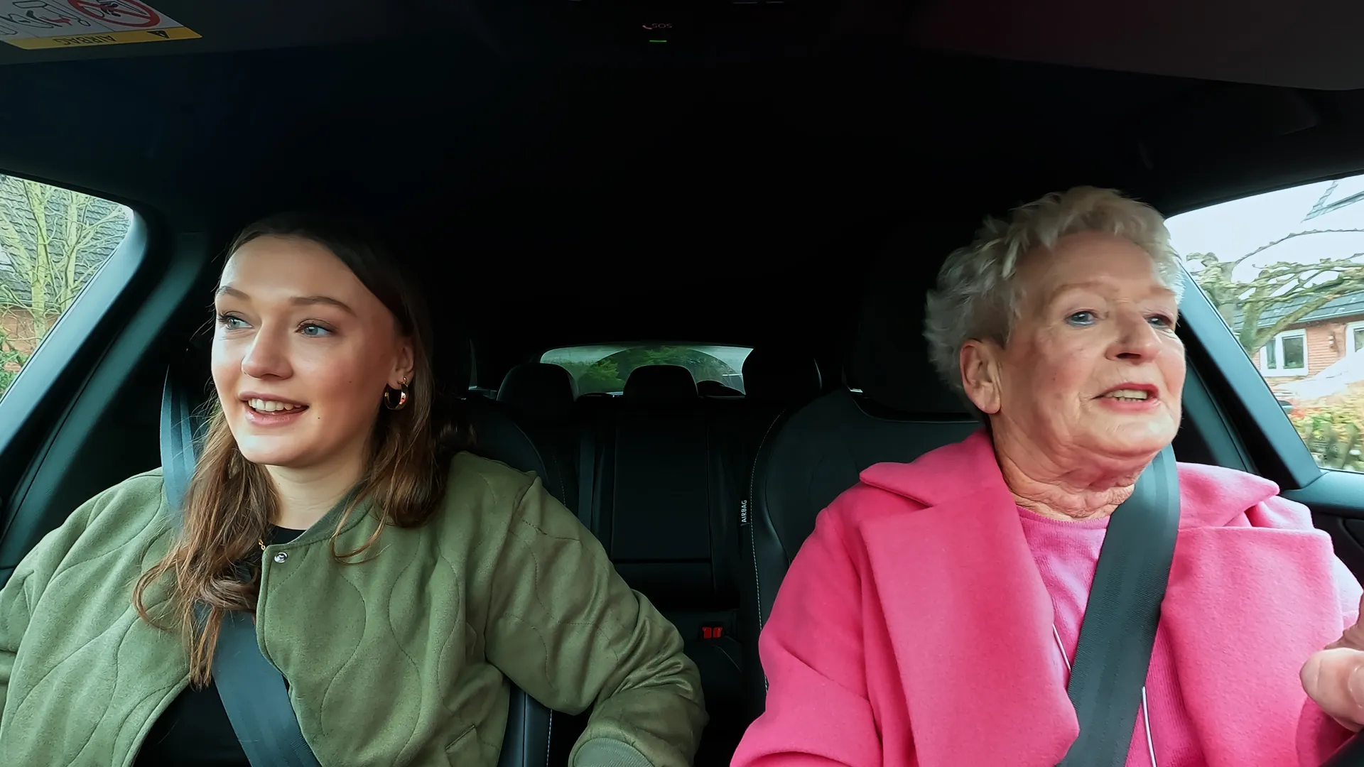 Eine ältere Frau in einem pinken Mantel und eine jüngere Frau in einer olivgrünen Jacke sitzen gemeinsam im Elektroauto. Es sind Oma Magrit und Enkelin Lilli. Magrit blickt konzentriert nach vorne, während Lilli lächelnd zur Seite blickt.