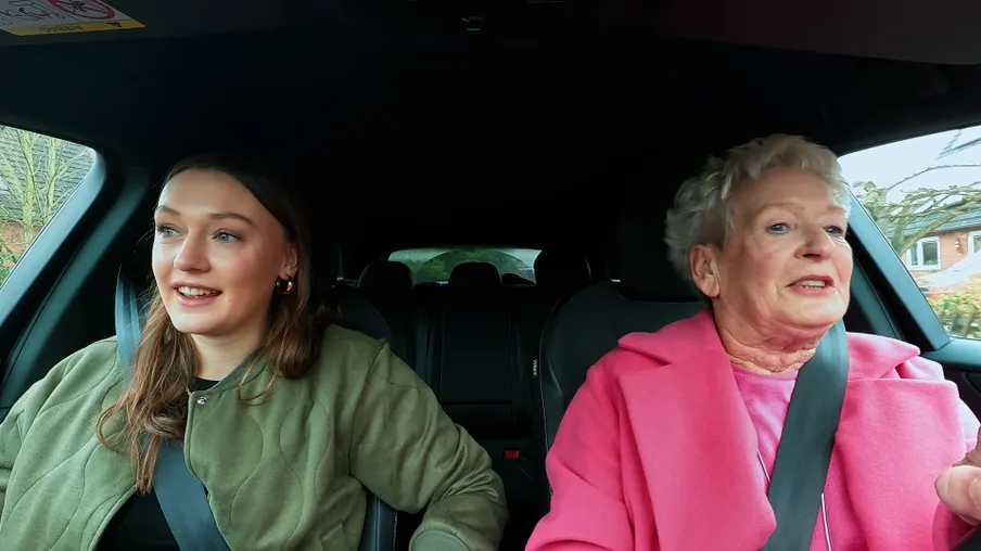 Eine ältere Frau in einem pinken Mantel und eine jüngere Frau in einer olivgrünen Jacke sitzen gemeinsam im Elektroauto. Es sind Oma Magrit und Enkelin Lilli. Magrit blickt konzentriert nach vorne, während Lilli lächelnd zur Seite blickt.