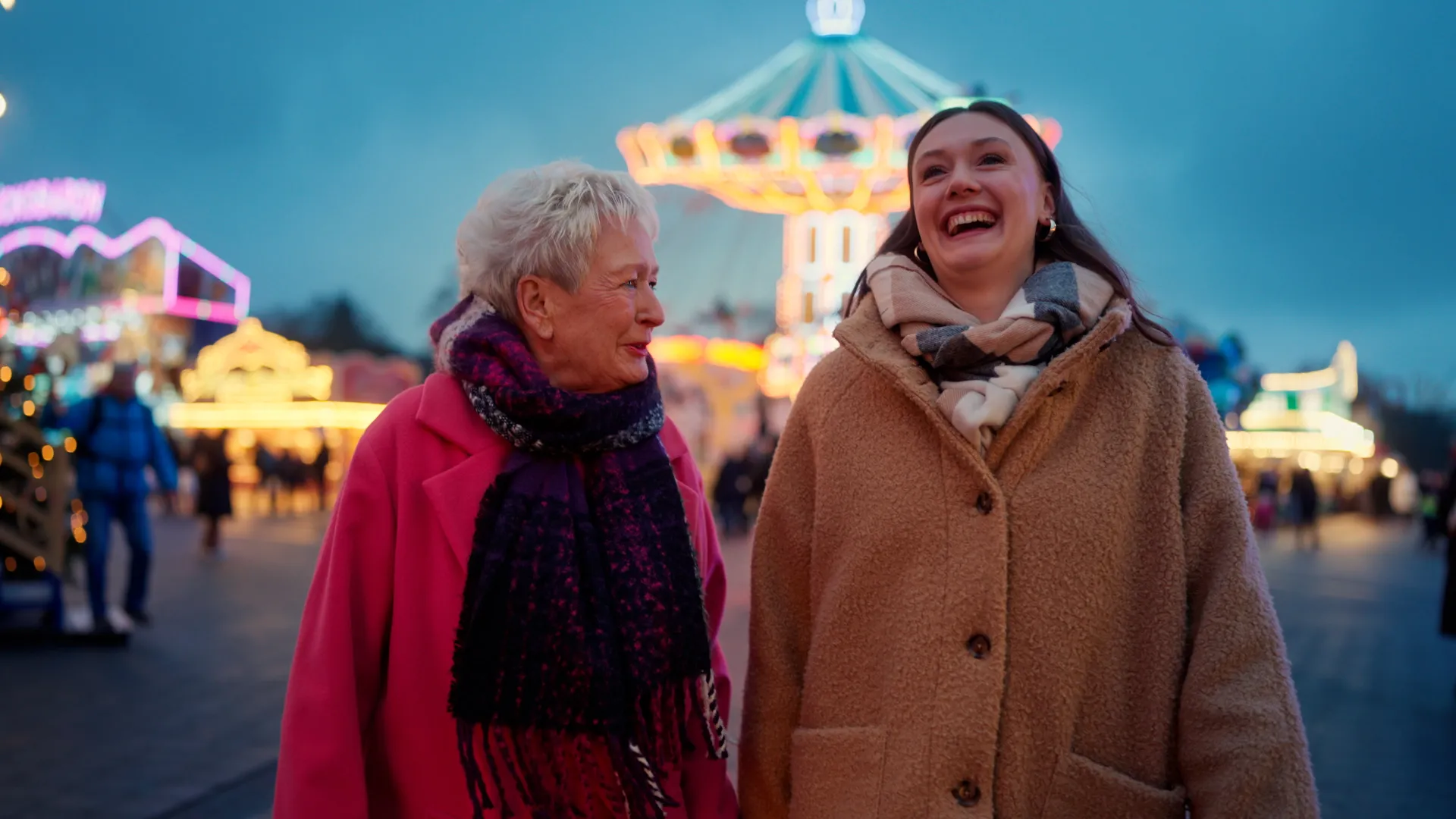 Eine ältere Frau in einem pinken Mantel und eine jüngere Frau in einem beigen Teddyfell-Mantel gehen lachend und sich anblickend über einen Jahrmarkt. Es sind Oma Magrit und Enkelin Lilli. Im Hintergrund sind Karussells und bunte Lichter zu sehen.