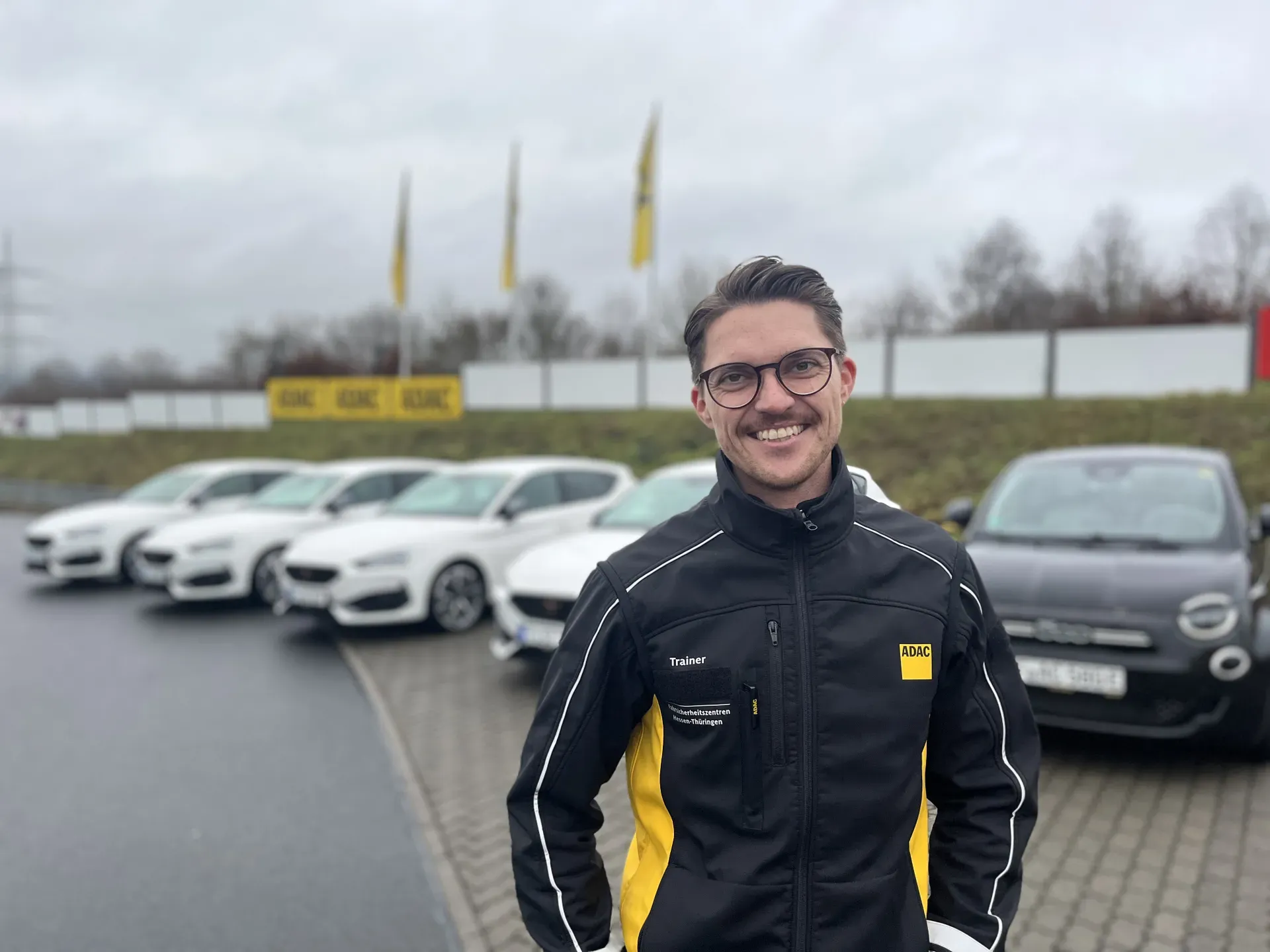 Ein lächelnder Mann in schwarz-gelber ADAC-Jacke steht auf einem Parkplatz vor einer Reihe geparkter Autos bei grauem Wetter. Es ist Lennart Schaub.