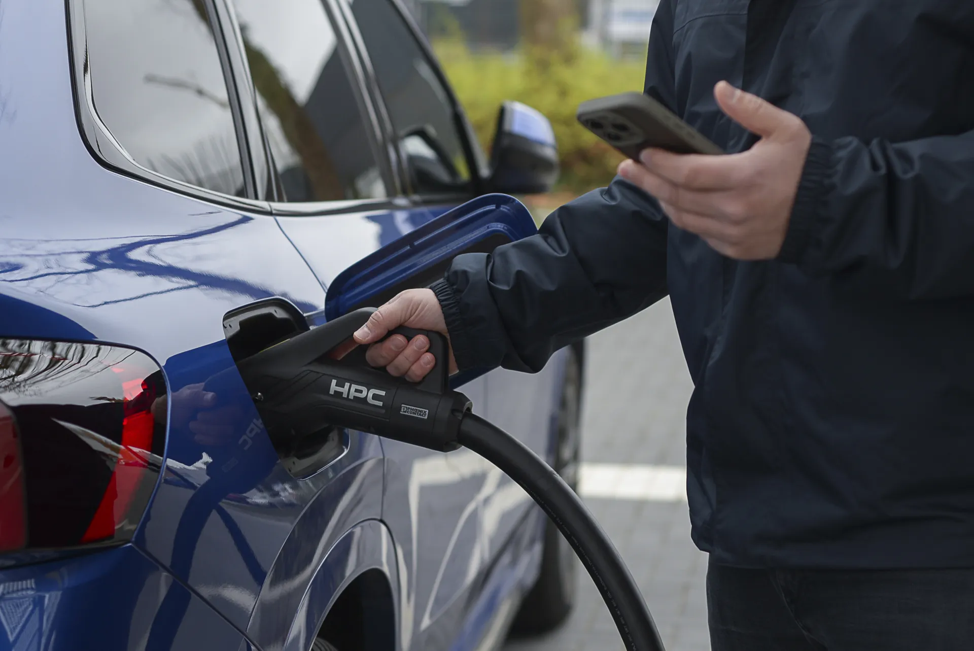 ine Person steckt einen Ladestecker in ein blaues E-Auto. In der anderen Hand hält sie ein Smartphone. Der Ladevorgang findet an einer Schnellladesäule statt.