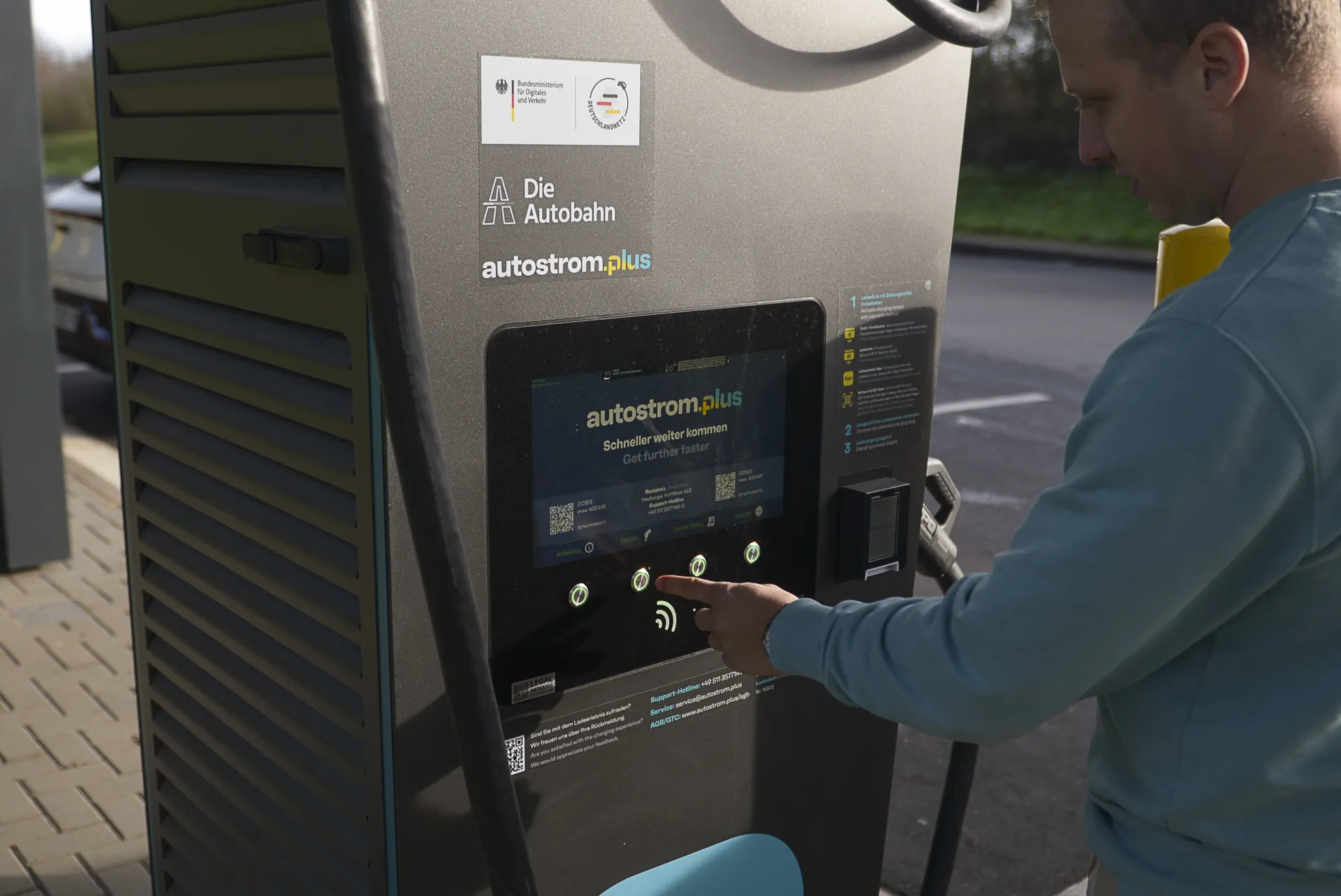 Ein Mann bedient das Touch-Display einer Deutschlandnetz- Ladesäule für E-Autos. Auf der Säule sind die Logos vom Bundesverkehrsministerium, vom Deutschlandnetz, von „Die Autobahn“ und autostrom.plus zu sehen.