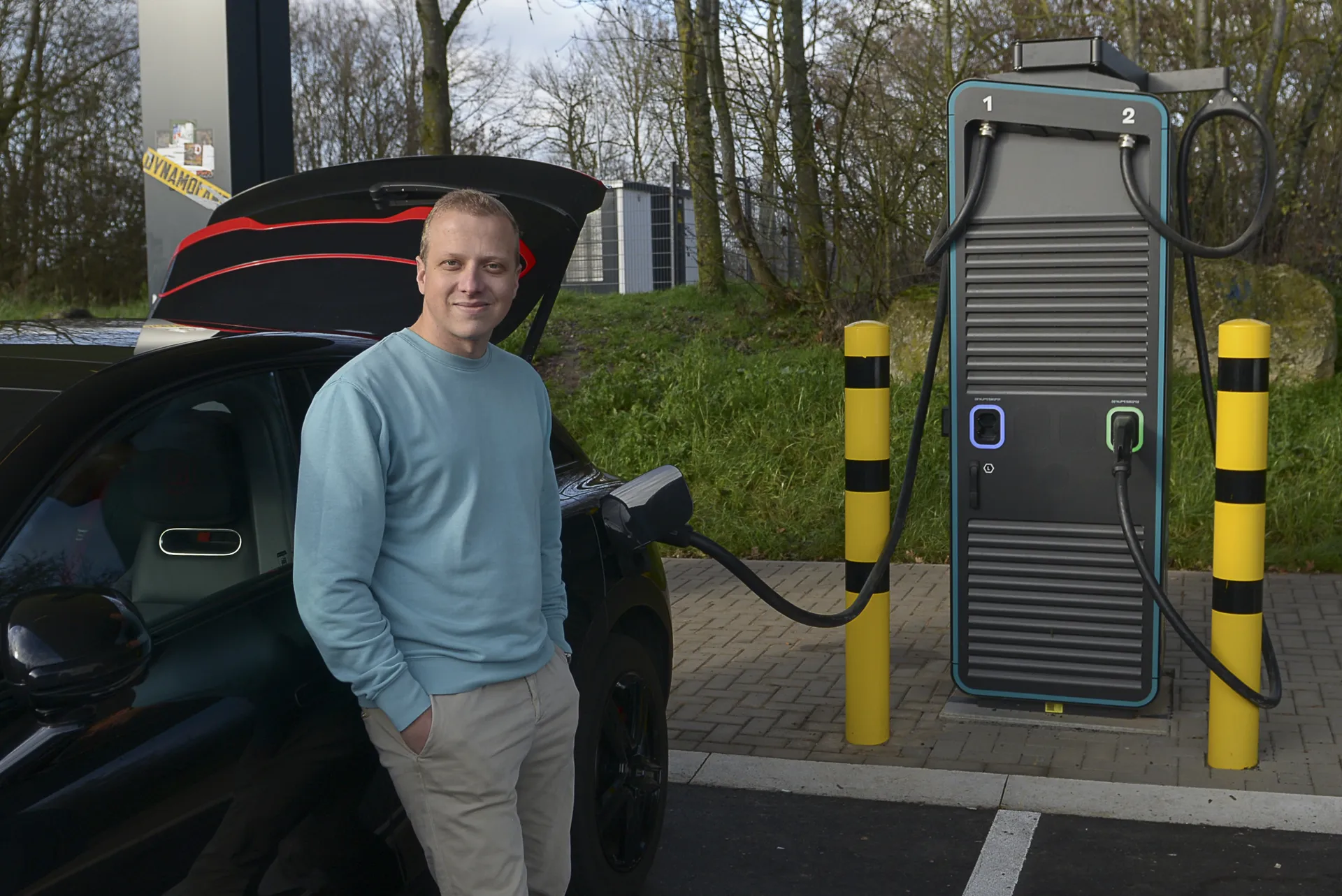 Ein Mann in einem türkisen Pullover lächelt in die Kamera, während sein schwarzes Elektroauto mit offenem Kofferraum an einer Schnellladesäule lädt.