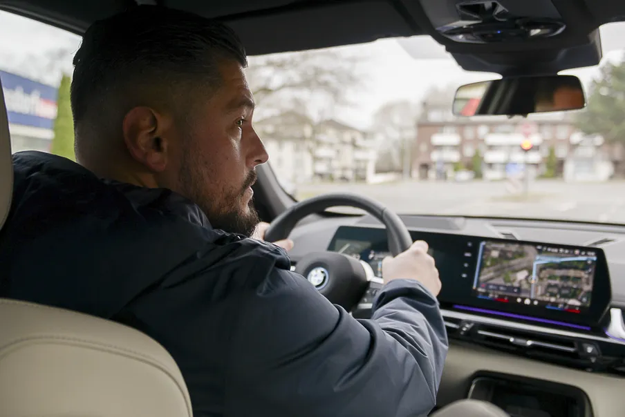 Ein Mann mit Bart sitzt am Steuer eines Elektroautos und blickt konzentriert auf die Straße. Im digitalen Cockpit läuft die Navigation auf einem großen Display.