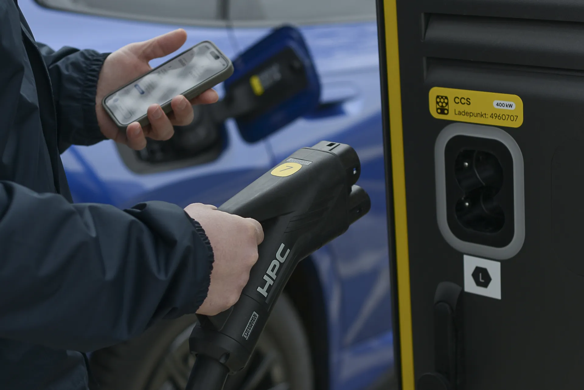 Nahaufnahme von Händen, die ein Smartphone und einen Ladestecker für E-Autos vor einer Schnellladesäule halten. Im Hintergrund ist ein blaues Elektroauto zu sehen.