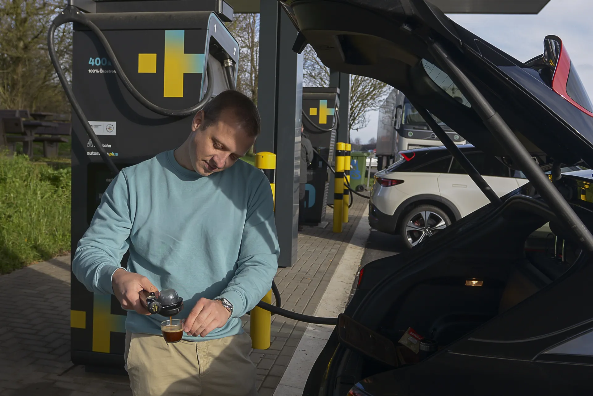 Ein Mann im türkisen Pullover gießt frischen Espresso aus einer Hand-Espressomaschine in eine Tasse, während sein schwarzes Elektroauto an einer Schnellladesäule lädt.