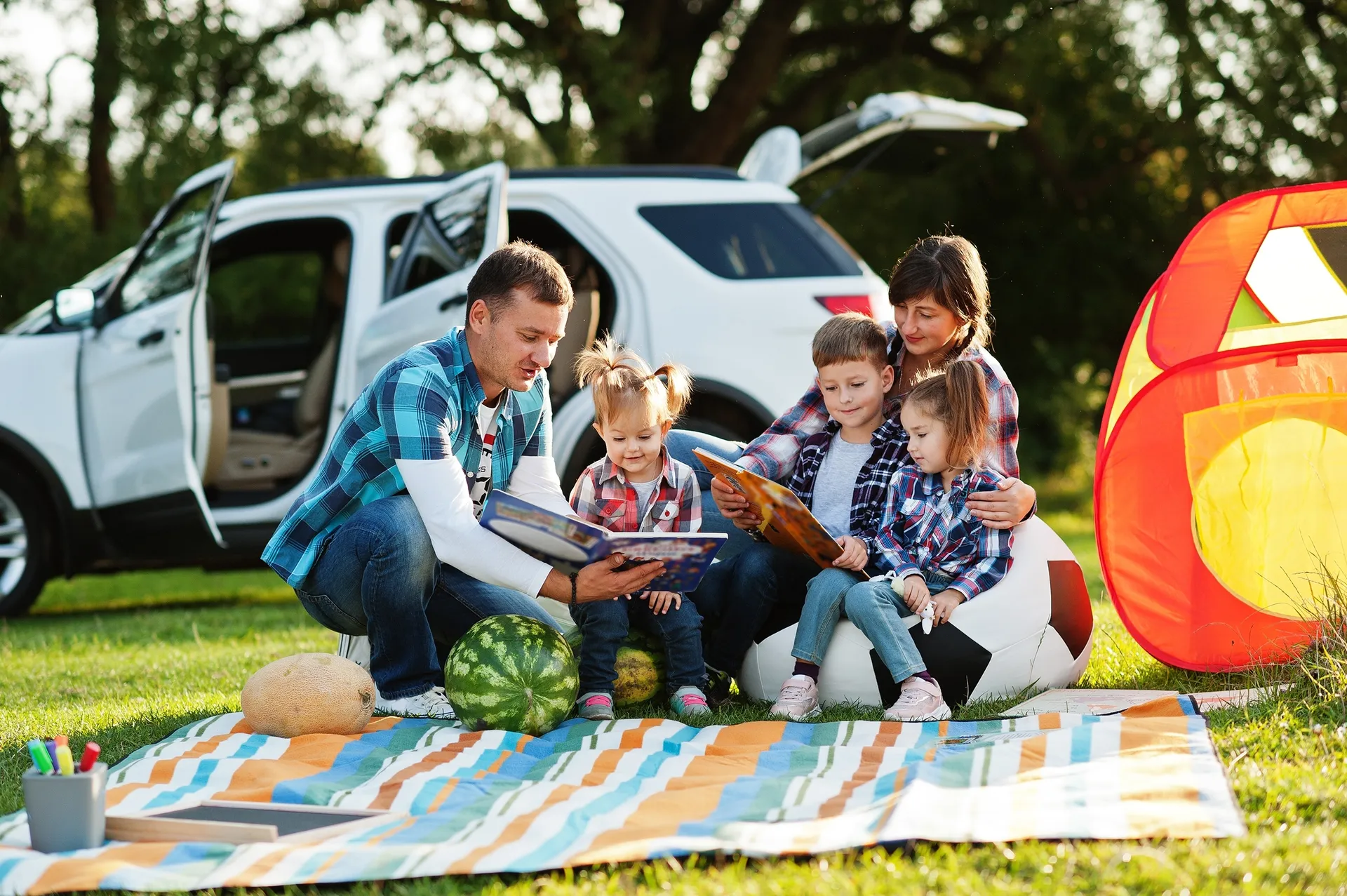 Eine Familie, bestehend aus Vater, Mutter und drei Kindern, sitzt auf einer bunten Picknickdecke im Gras. Im Hintergrund steht ein weißes Auto mit geöffneten Türen.