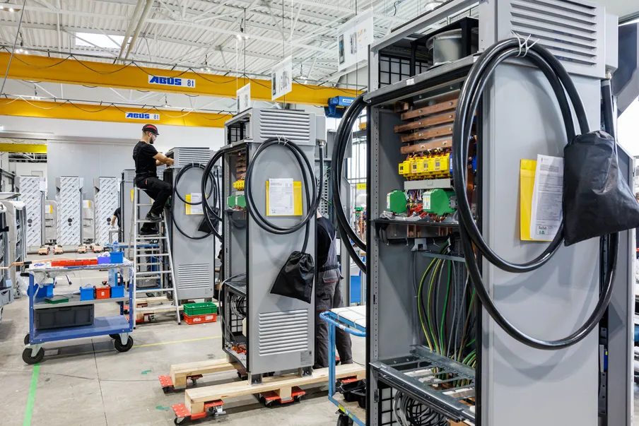Mitarbeiter arbeiten in einer Fabrikhalle an der Montage großer, grauer Schaltschränke mit sichtbaren Kabeln und elektrischen Komponenten.