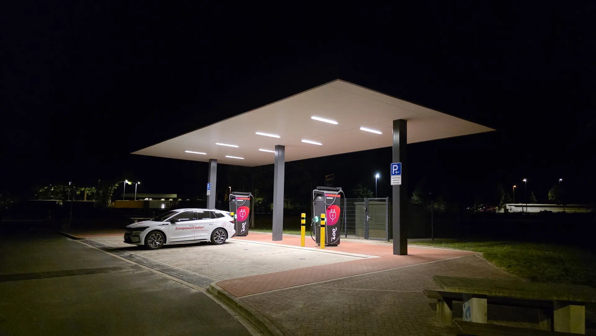 Nachtaufnahme eines überdachten Deutschlandnetz-Ladeparks. Ein weißer Kombi parkt an einer von zwei Ladesäulen.