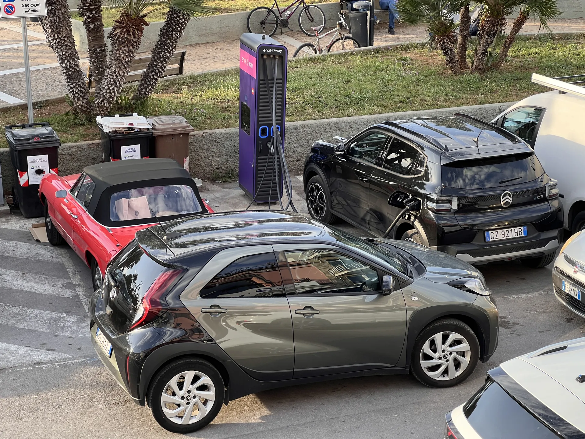 Ein schwarzes Elektroauto lädt an einer lila Ladesäule auf einem Parkplatz. Daneben parkt ein rotes Cabrio, davor ein graues Auto.