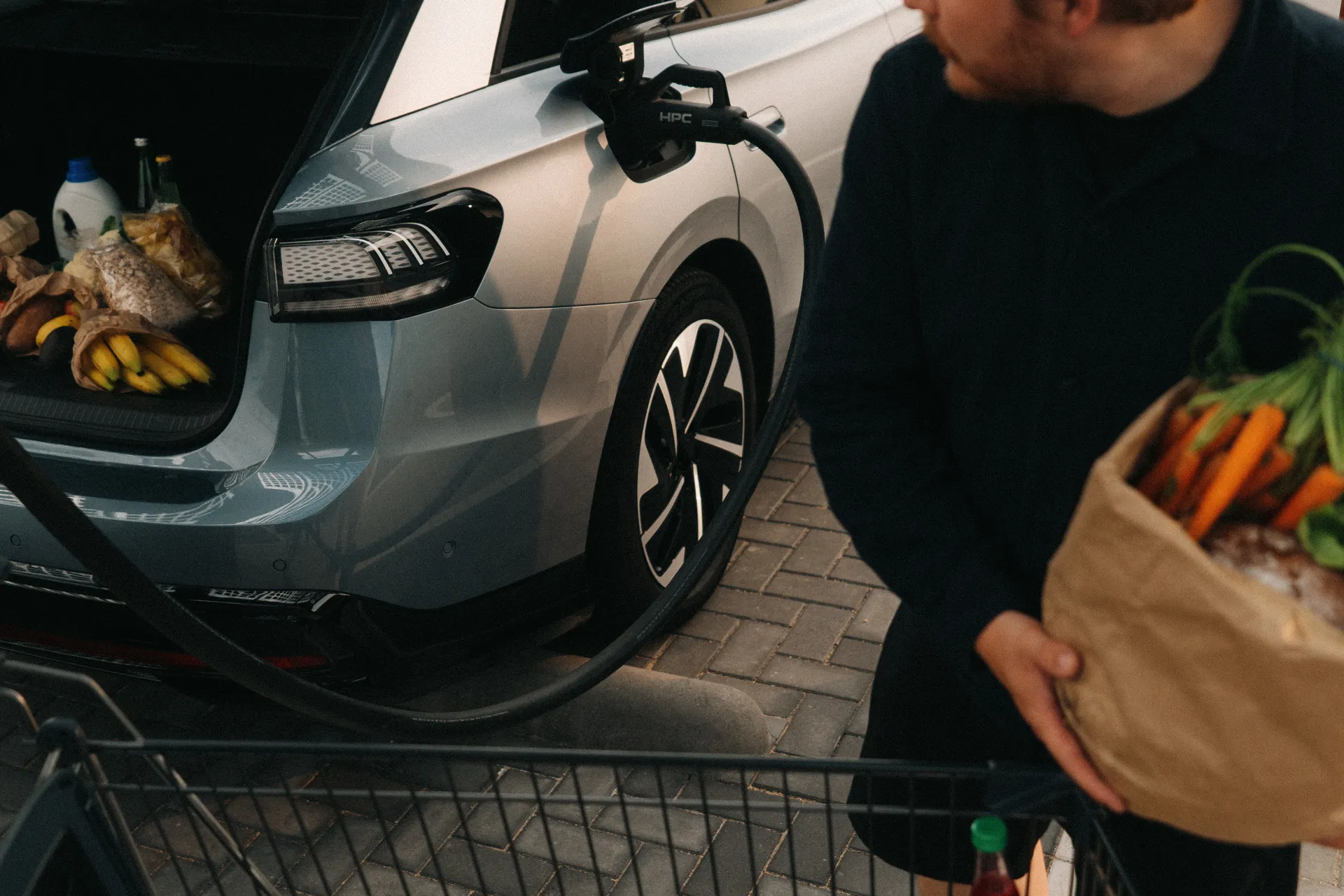Graues E-Auto mit angeschlossenem Ladekabel und geöffnetem Kofferraum voller Einkäufe. Eine Person trägt eine Papiertüte mit Karotten und Gemüse. Einkaufswagen im Vordergrund.