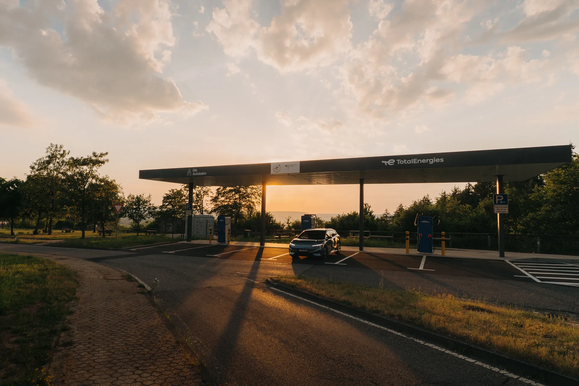 Schnellladestation mit Überdachung auf einem Autobahnrastplatz bei Sonnenuntergang. Ein E-Auto steht an einer Ladesäule. Bäume und Parkplatzmarkierungen sind sichtbar.