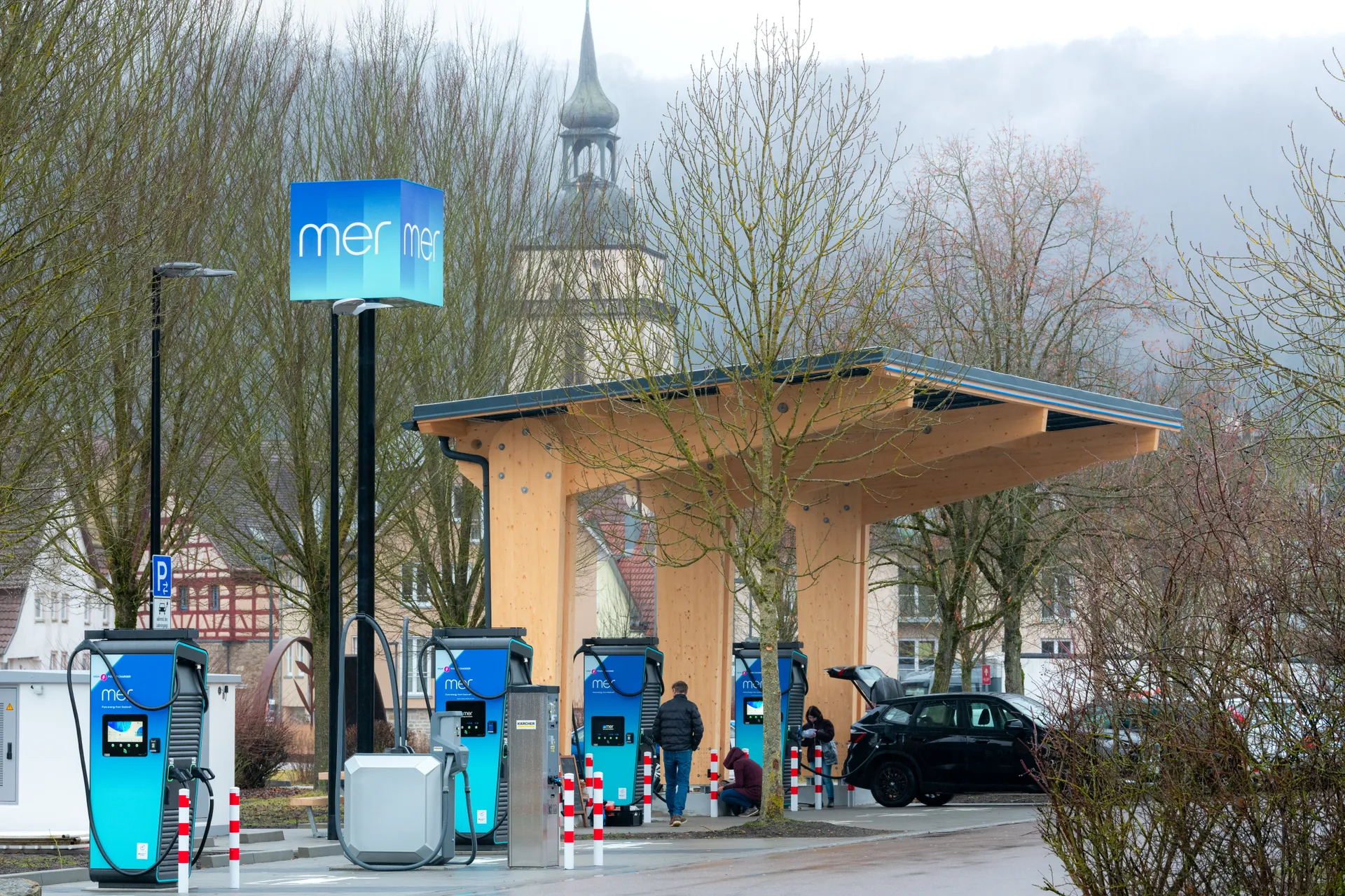 Mehrere blaue Schnellladesäulen vom Deutschlandnetz unter einem Holzdach. Ein schwarzes Elektroauto lädt, Personen stehen daneben. Im Hintergrund ist ein Kirchturm zu erkennen.