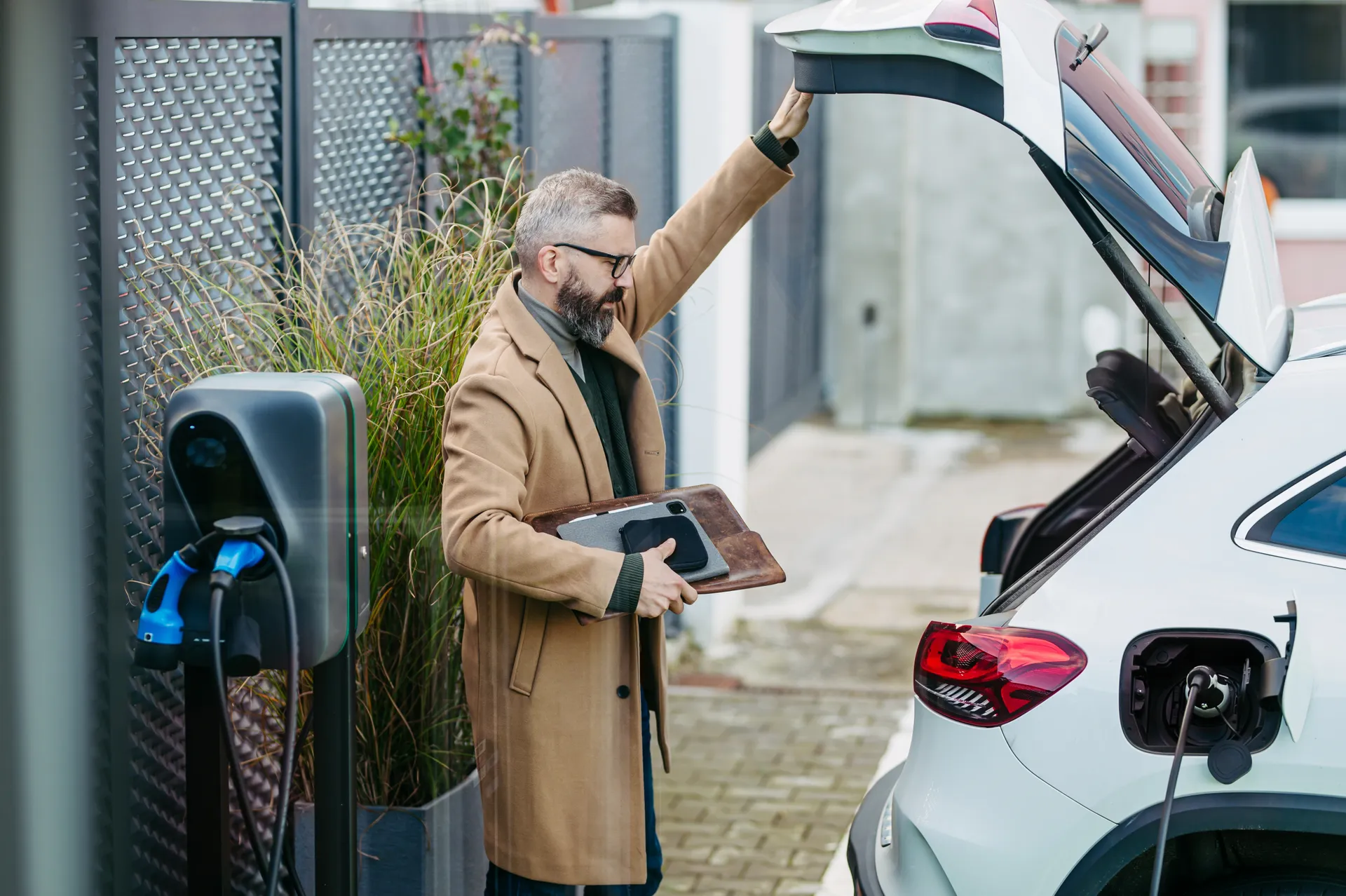 Ein Mann mit Brille im beigen Mantel steht neben einem weißen E-Auto, das an einer Ladesäule lädt. Er öffnet die Heckklappe und hält Unterlagen in der Hand.