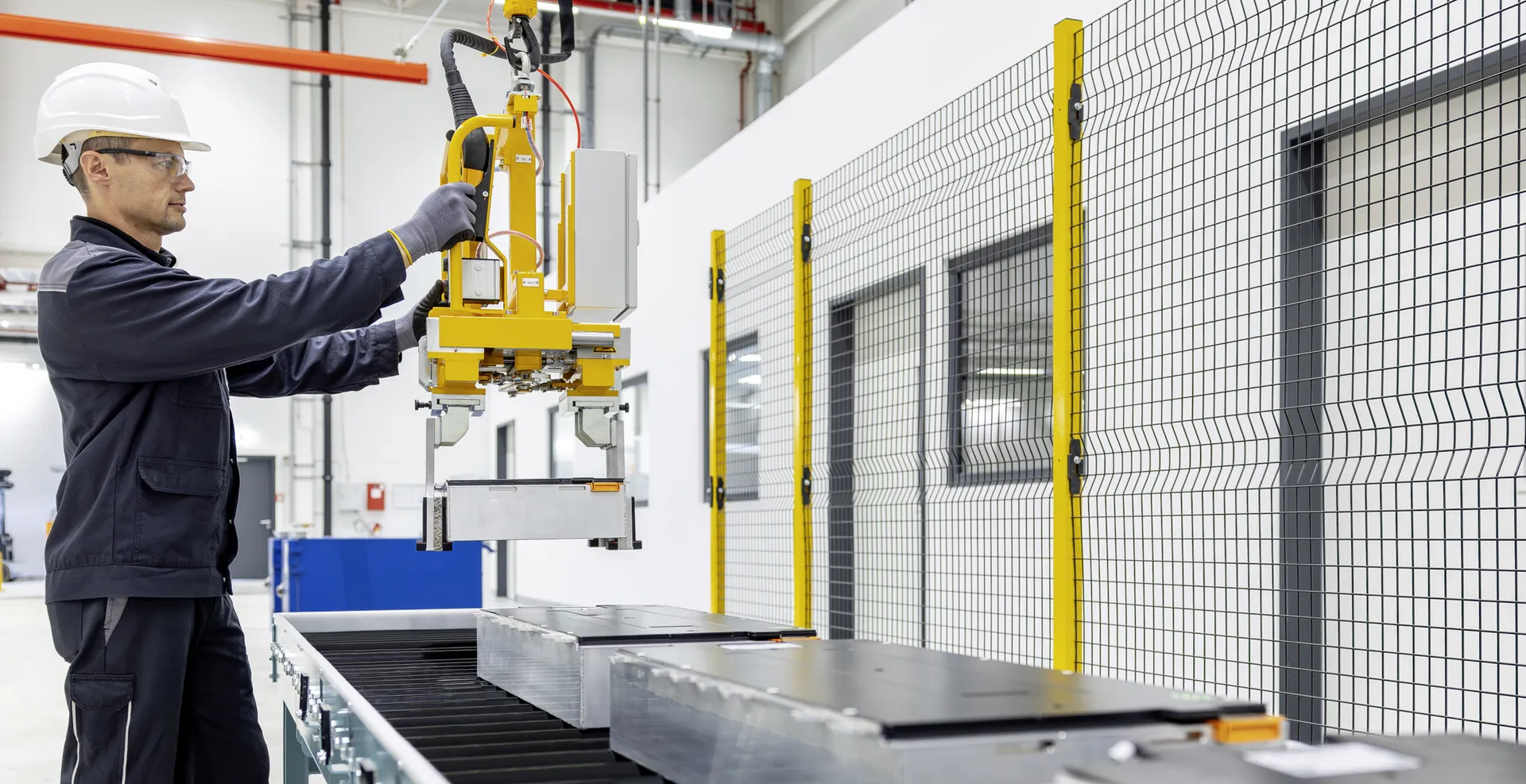 Ein Arbeiter in weißem Schutzhelm und dunkler Arbeitskleidung bedient einen gelben Kran, der Batteriemodule auf ein Förderband in einer Batterierecyclinganlage hebt.