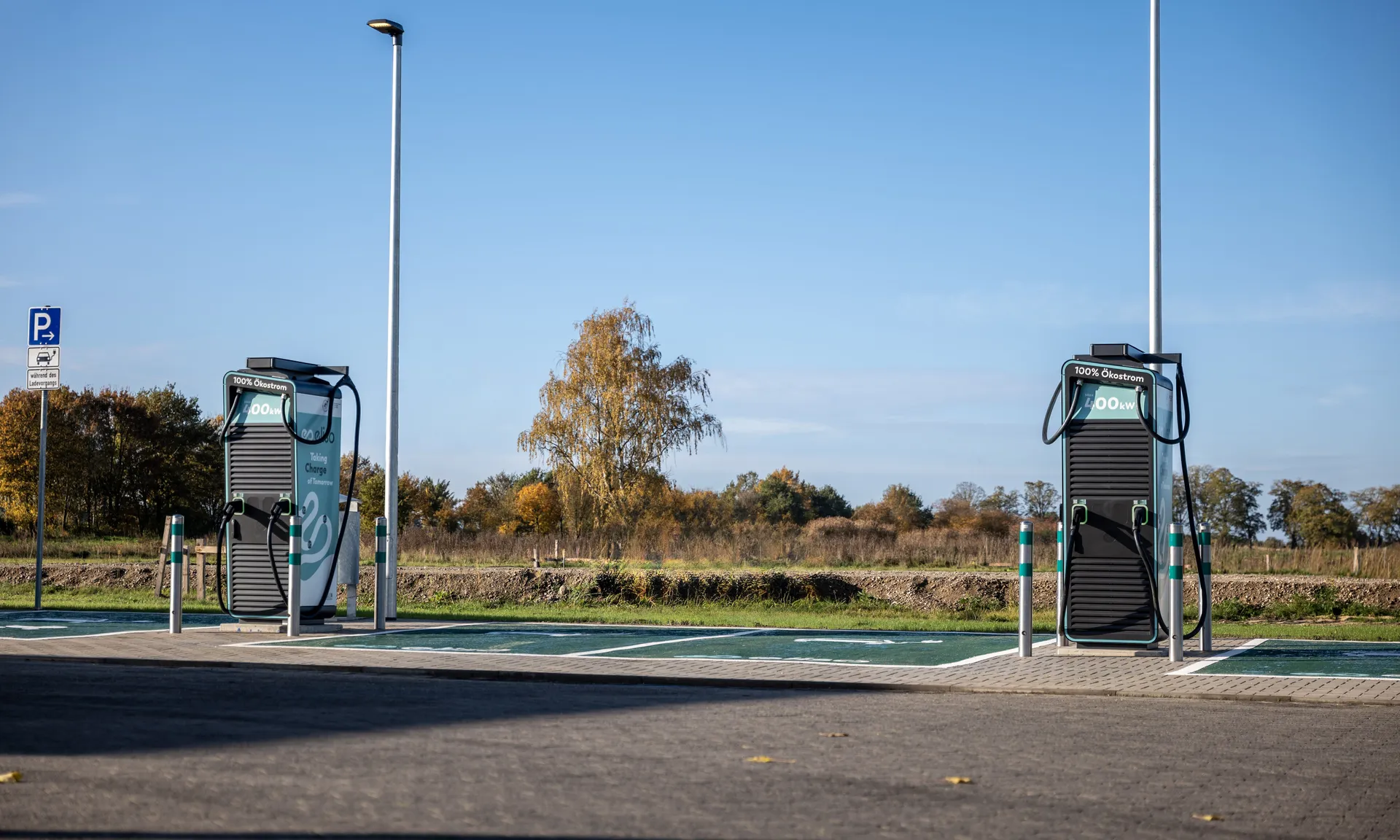 Zwei Deutschlandnetz-Ladesäulen für Elektroautos stehen auf einem Parkplatz mit grün markierten Stellflächen. Im Hintergrund sind Bäume und ein Feld unter strahlend blauem Himmel zu sehen.