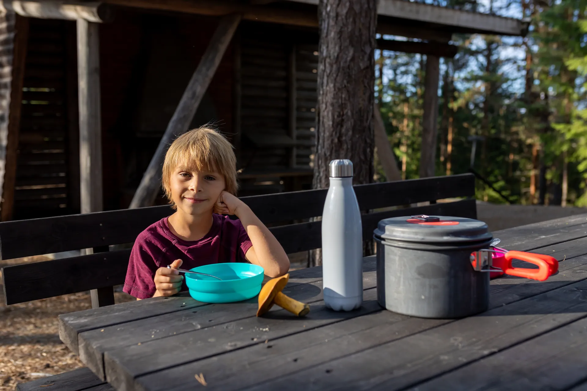 Ein kleiner Junge mit blonden Haaren sitzt lächelnd an einem rustikalen Holztisch. Auf dem Tisch stehen Campinggeschirr und ein frisch gesammelter Pilz.