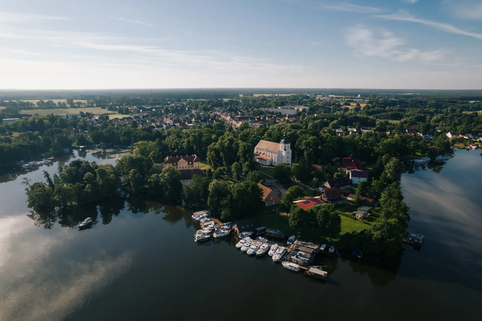 Luftbild einer grünen Ortschaft am Seeufer mit Bootsanleger. Es ist Mirow. Eine weiße Kirche mit zwei Türmen ragt heraus. Wohnhäuser und Bäume umgeben den Hafen.