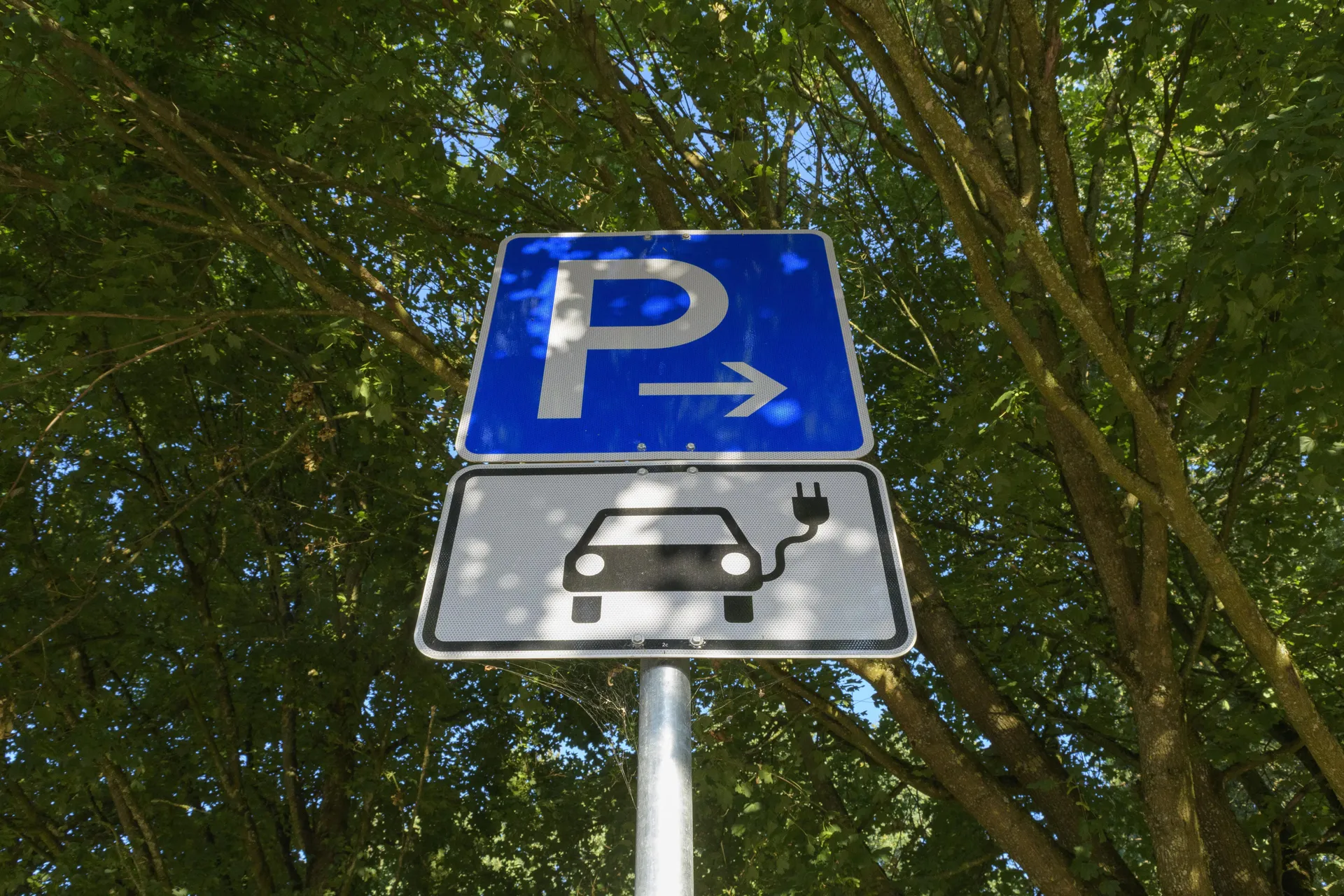 Nahaufnahme eines blauen Parkplatzschildes mit weißem P und Pfeil nach rechts. Darunter ein weißes Zusatzzeichen mit dem Piktogramm eines Elektroautos.