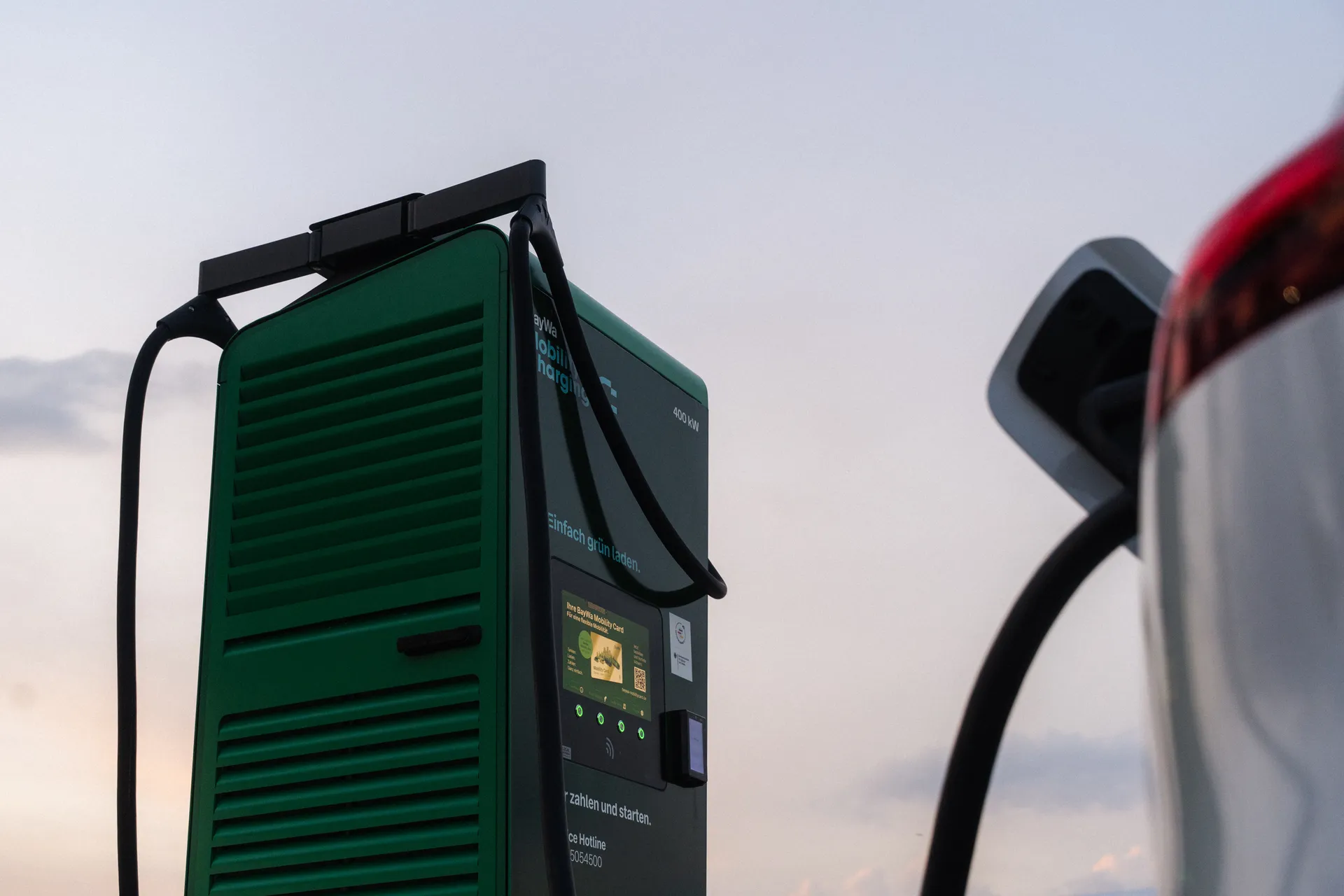 Nahaufnahme einer grünen Ladesäule mit Touchscreen und Ladekabeln. Aufschrift: „400 kW" und „Einfach grün laden." Rechts ein silbernes E-Auto.