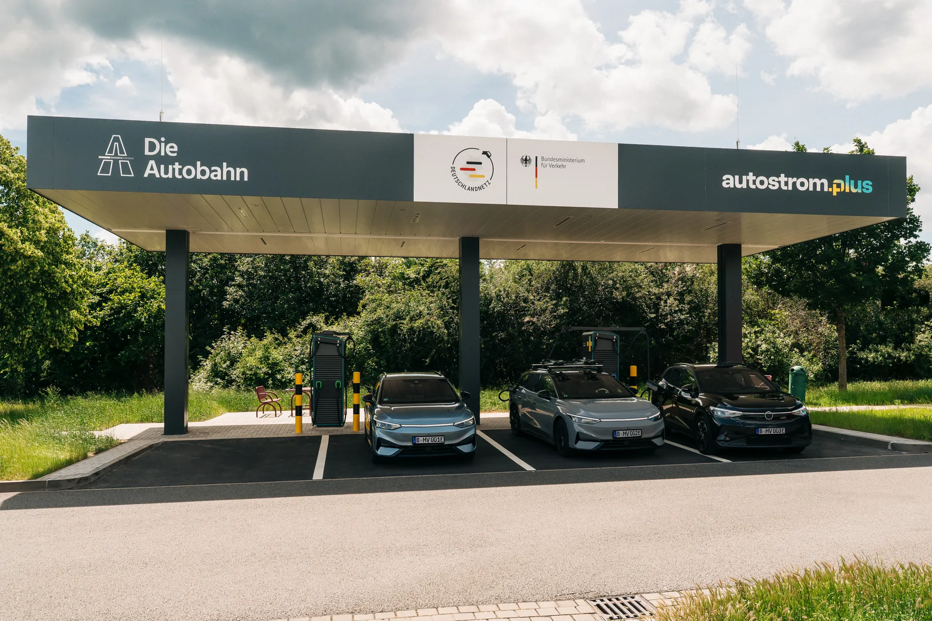 Überdachte Schnellladestation mit drei E-Autos an Ladesäulen. Auf dem Dach sind die Logos von Die Autobahn GmbH, autostrom.plus, Deutschlandnetz und Bundesministerium für Verkehr zu sehen.