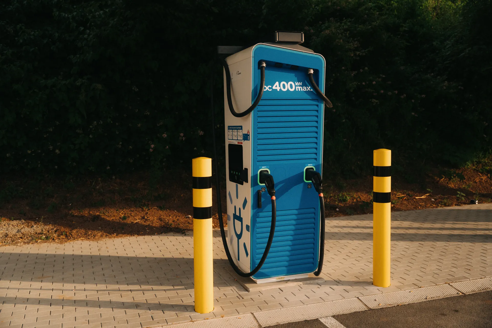 Blau-weiße Schnellladesäule von mit der Aufschrift DC 400 kW max und zwei Ladekabeln. Gelb-schwarze Poller stehen daneben.