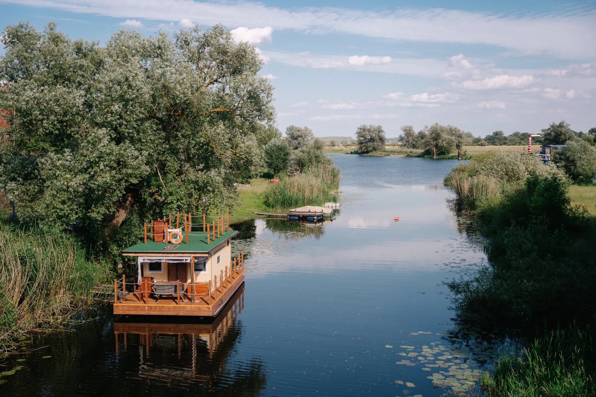 Hausboot mit grünem Dach und Holzterrasse auf einem stillen Fluss. Ein Rettungsring hängt an Bord. Weiden, Schilf und Seerosen prägen die naturbelassene Umgebung.