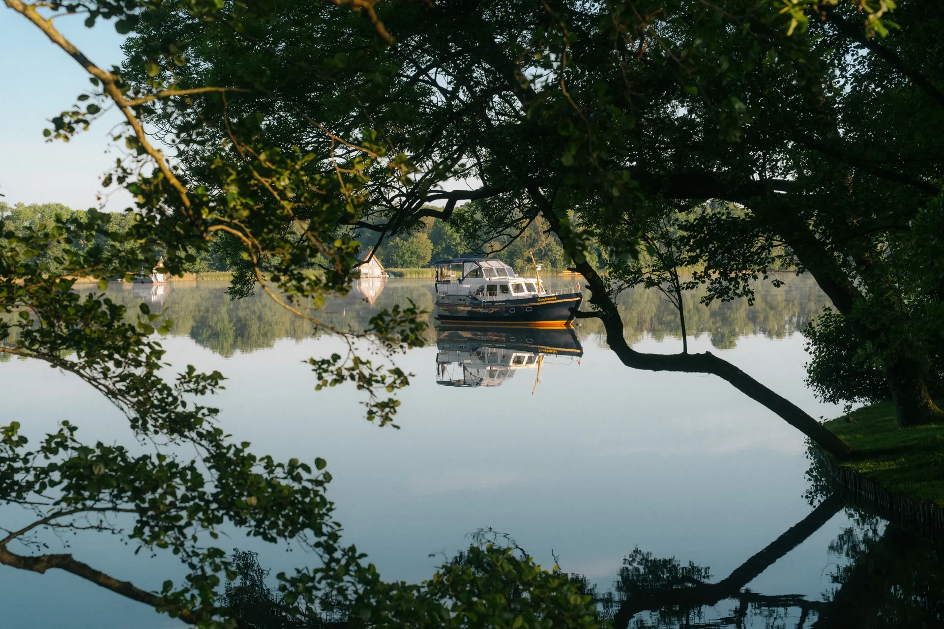 Motorboot mit dunnklem Rumpf und gelbem Streifen auf einem stillen See. Das Boot spiegelt sich im Wasser. Äste im Vordergrund rahmen den Blick ein.