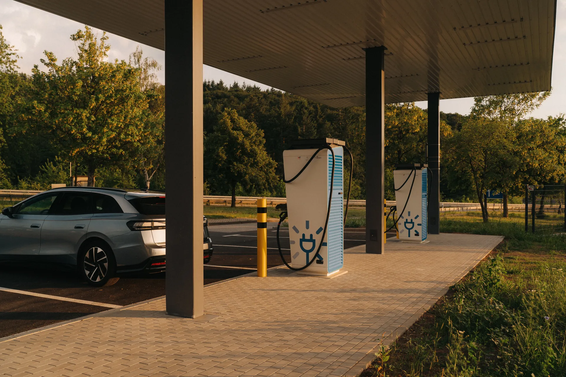 Überdachte Ladestation mit zwei weißen Schnellladesäulen und blauem Steckersymbol. Ein graues E-Auto steht links im Bild. Bäume und Wiese im Hintergrund.