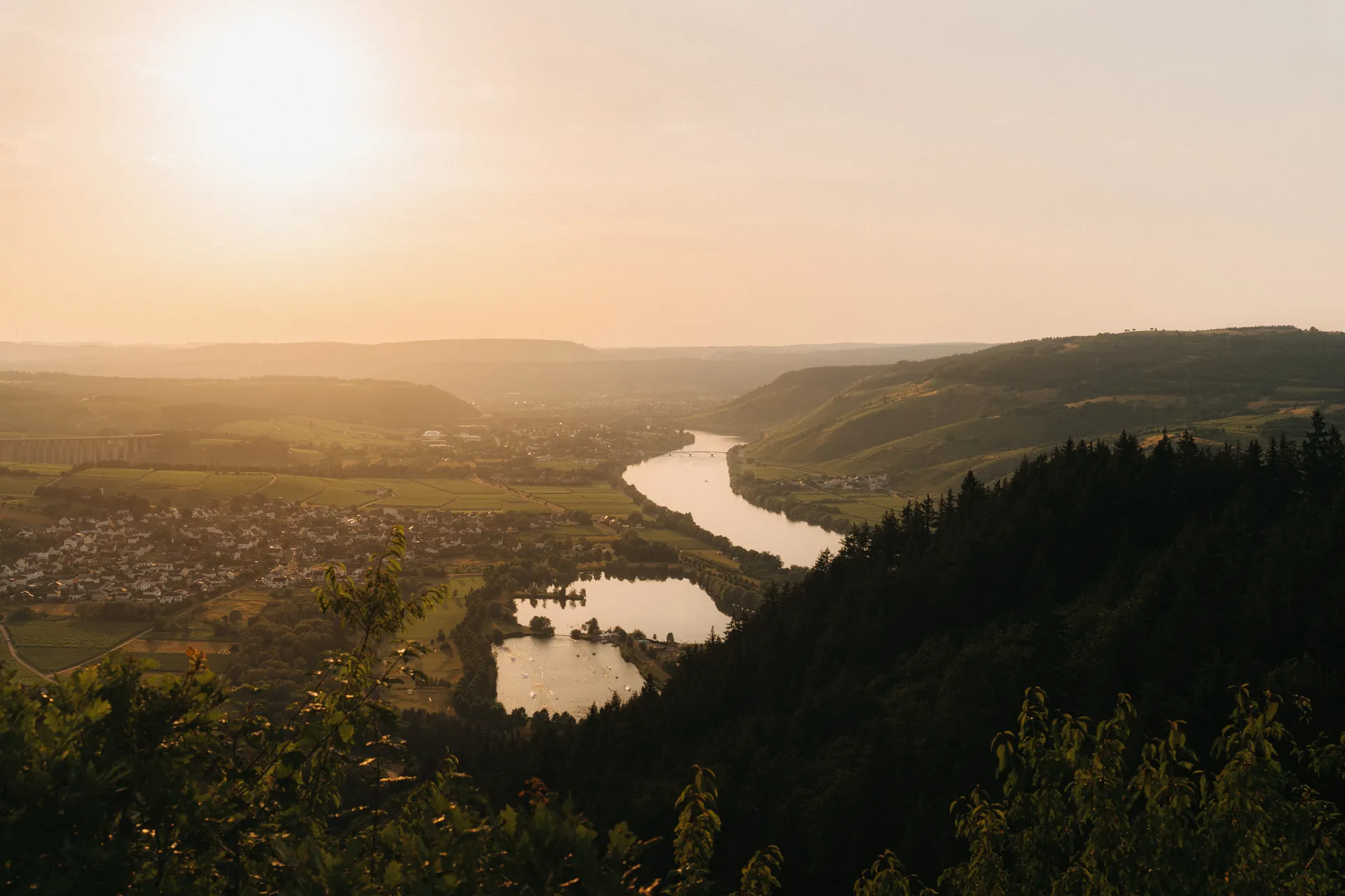 Weiter Blick über ein Flusstal im Abendlicht. Der Fluss windet sich durch grüne Hügel und Wälder. Eine Ortschaft liegt am linken Ufer, eine Brücke überspannt den Fluss.