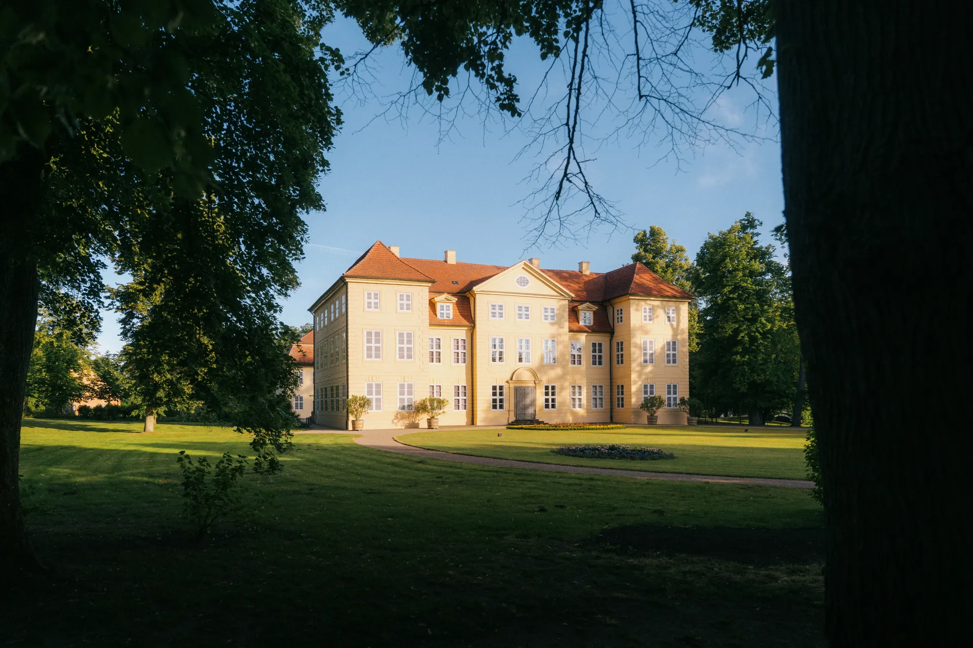 Herrenhaus mit hellgelber Fassade und rotem Ziegeldach. Davor liegt eine Rasenfläche mit Blumenbeet. Bäume säumen den Schlosspark.