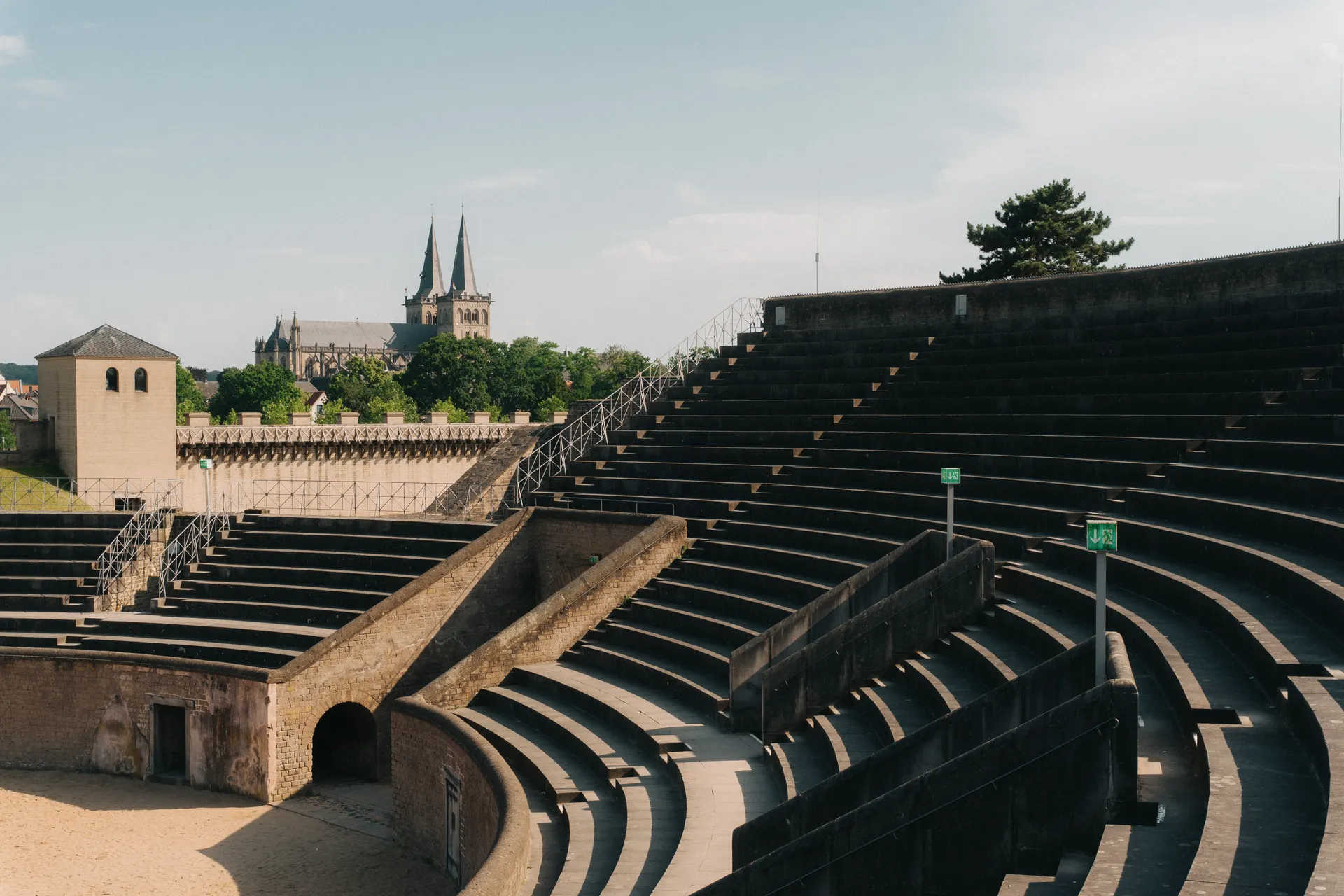 Amphitheater mit halbrunden Steintribünen und Treppen. Links steht ein historischer Turm. Im Hintergrund eine Kirche mit zwei Türmen.
