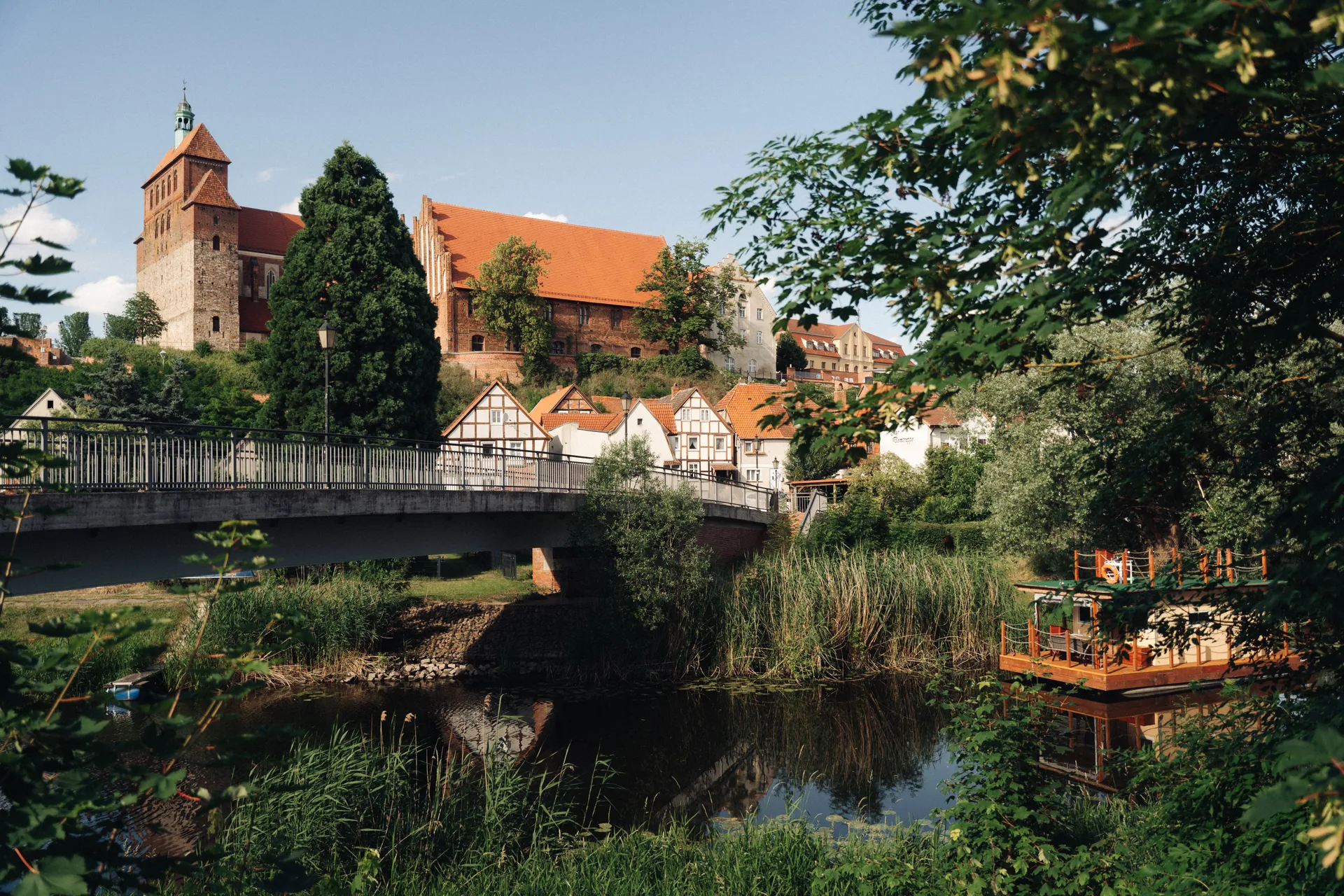 Historische Altstadt mit Fachwerkhäusern am Flussufer. Eine Fußgängerbrücke führt in die Stadt. Im Hintergrund eine Kirche mit Turm und roten Dächern.