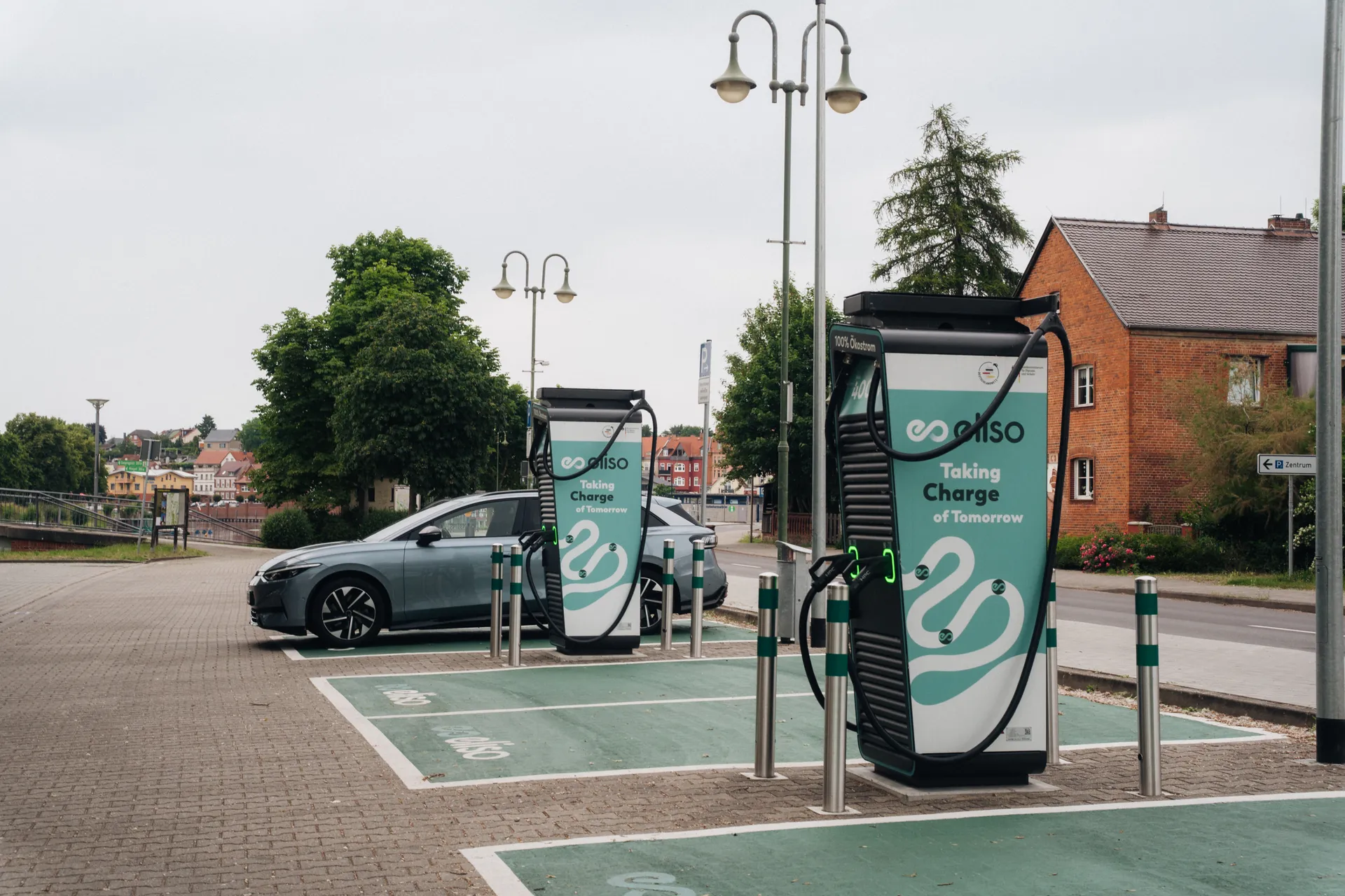 Graublaues E-Auto an einer Schnellladesäule mit türkis-weißem Design von dem Betreiber eliso. Mehrere Ladepunkte stehen auf markierten Parkflächen. Im Hintergrund ein Backsteinhaus.