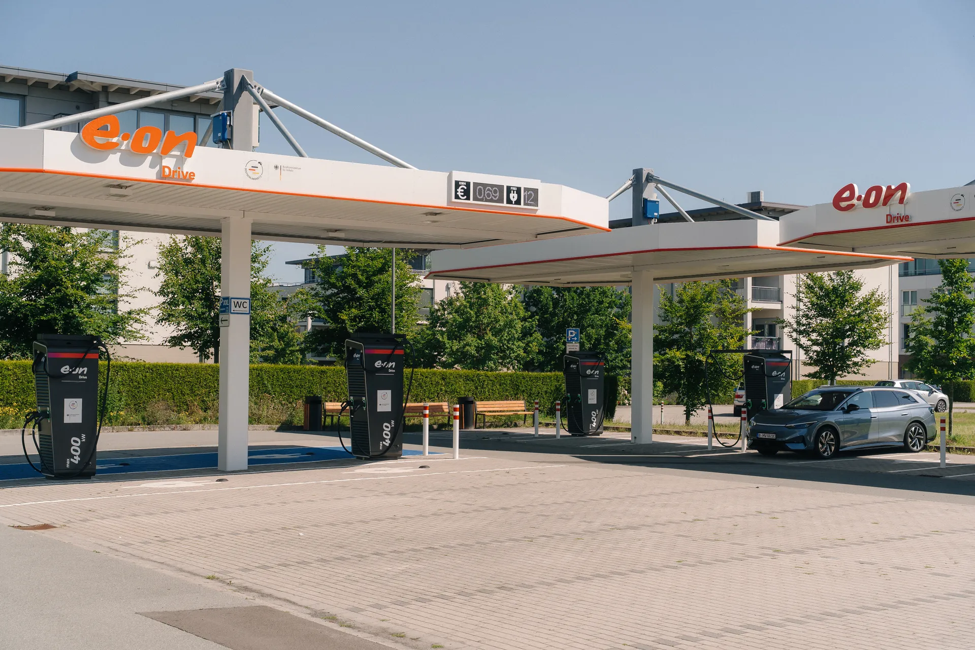 Überdachte Schnellladestation mit mehreren schwarzen Ladesäulen. Ein graues E-Auto lädt an einer Säule. Eine digitale Anzeige zeigt den Strompreis von 0,69 Euro.