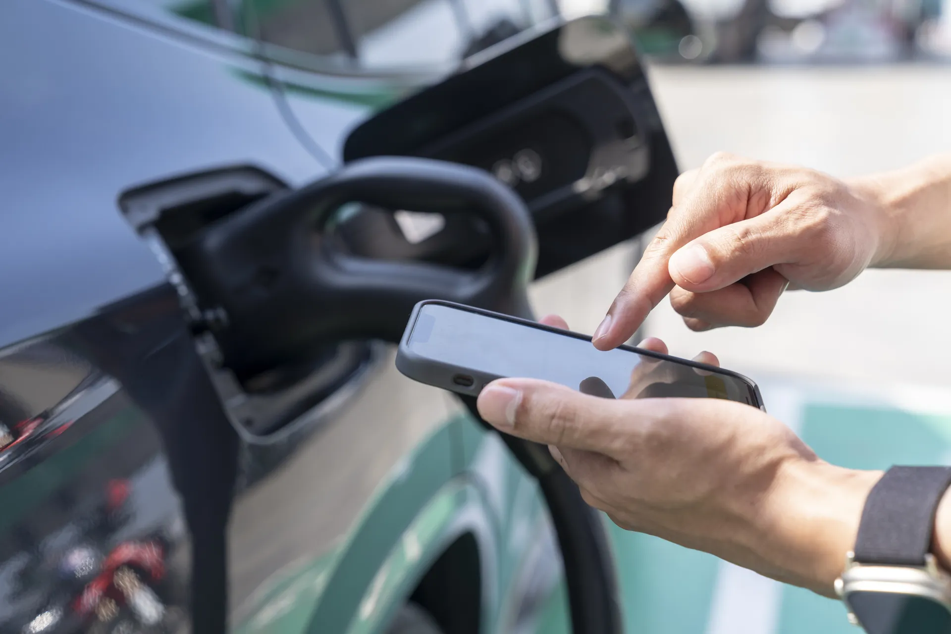 Nahaufnahme von zwei Händen, die ein Smartphone halten und den Bildschirm bedienen. Im unscharfen Hintergrund ist ein dunkles E-Auto zu sehen, in dessen geöffneter Ladeklappe ein schwarzes Ladekabel steckt.