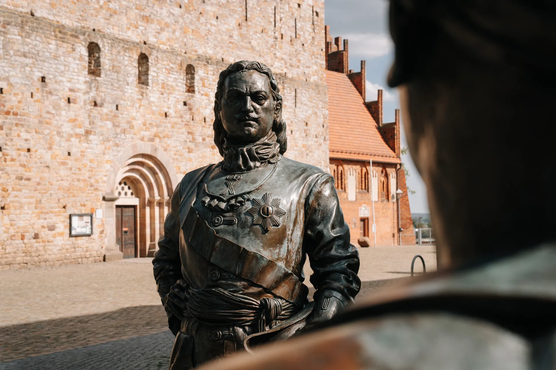 Bronzefigur eines Mannes in barocker Uniform mit Ordenskreuz. Im Hintergrund eine Steinmauer.