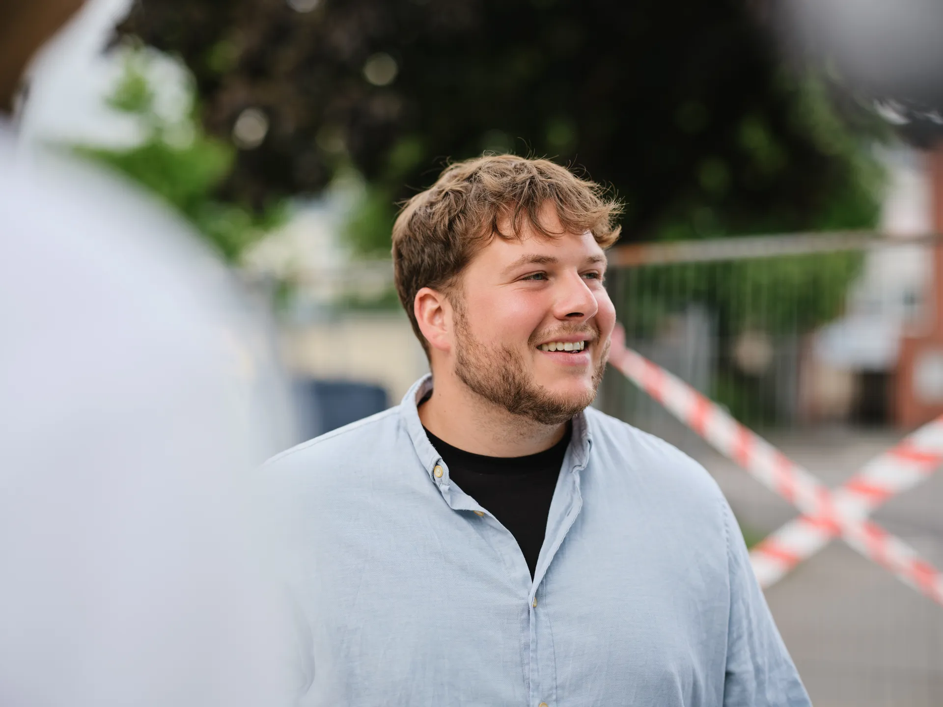 Nahaufnahme von Gideon Harzer, ein junger Mann mit kurzen, braunen Haaren und Bart, der freundlich lächelnd nach rechts blickt. Er trägt ein hellblaues Hemd über einem schwarzen T-Shirt. Der Hintergrund ist unscharf.