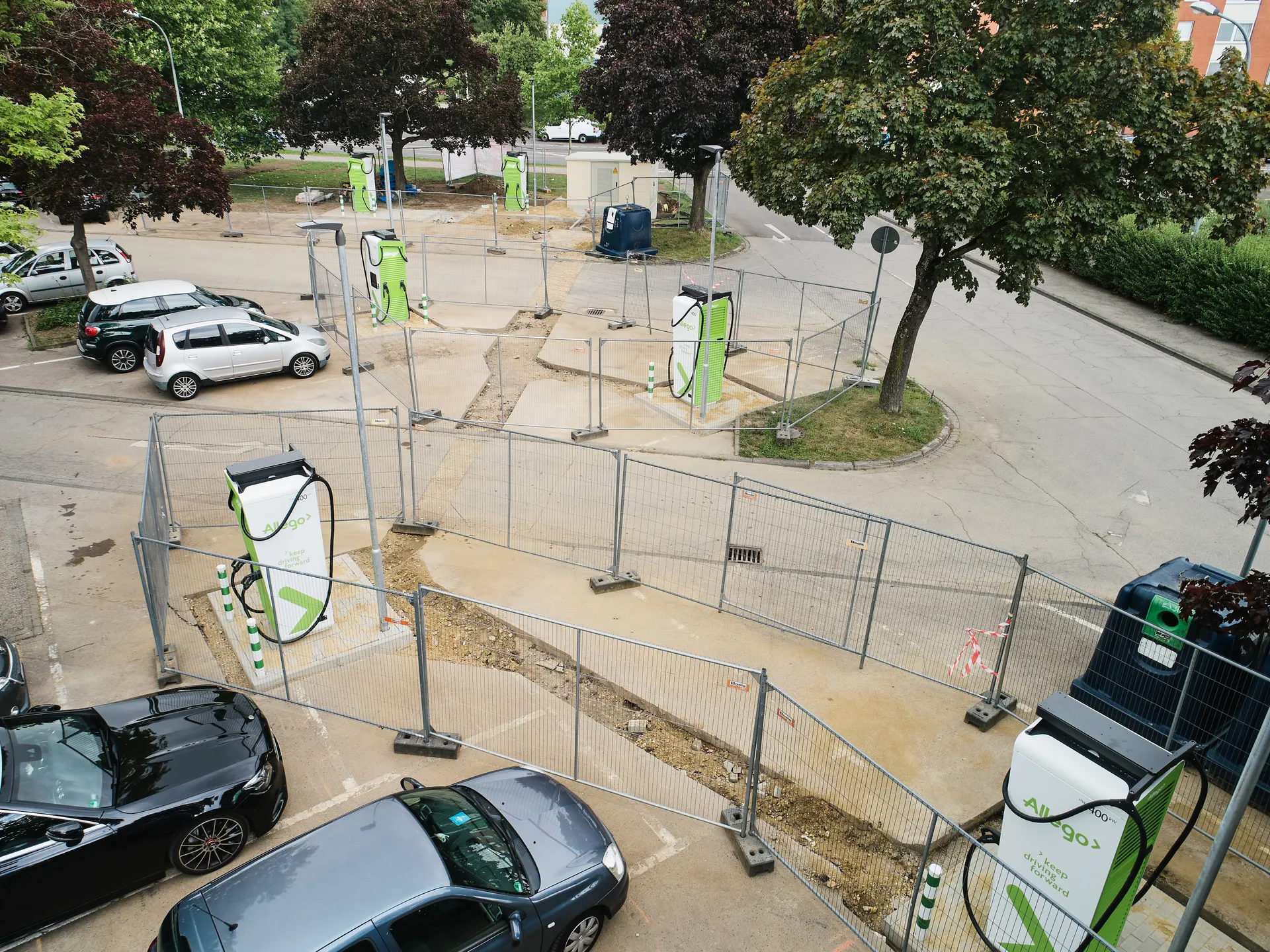Vogelperspektive auf die Baustelle für den neuen Ladepark in Eschweiler. Mehrere grün-weiße Ladesäulen der Marke Allego stehen auf drei Parkinseln und sind von Bauzäunen umgeben. Auf dem Parkplatz stehen einige Autos zwischen Bäumen.