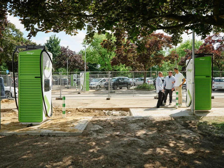 Drei Männer im Gespräch neben zwei neuen, grünen Schnellladesäulen von Allego auf einer Baustelle. Im Hintergrund ist durch einen Bauzaun ein Parkplatz mit mehreren Autos zu erkennen. Die Szene ist von Bäumen umgeben.