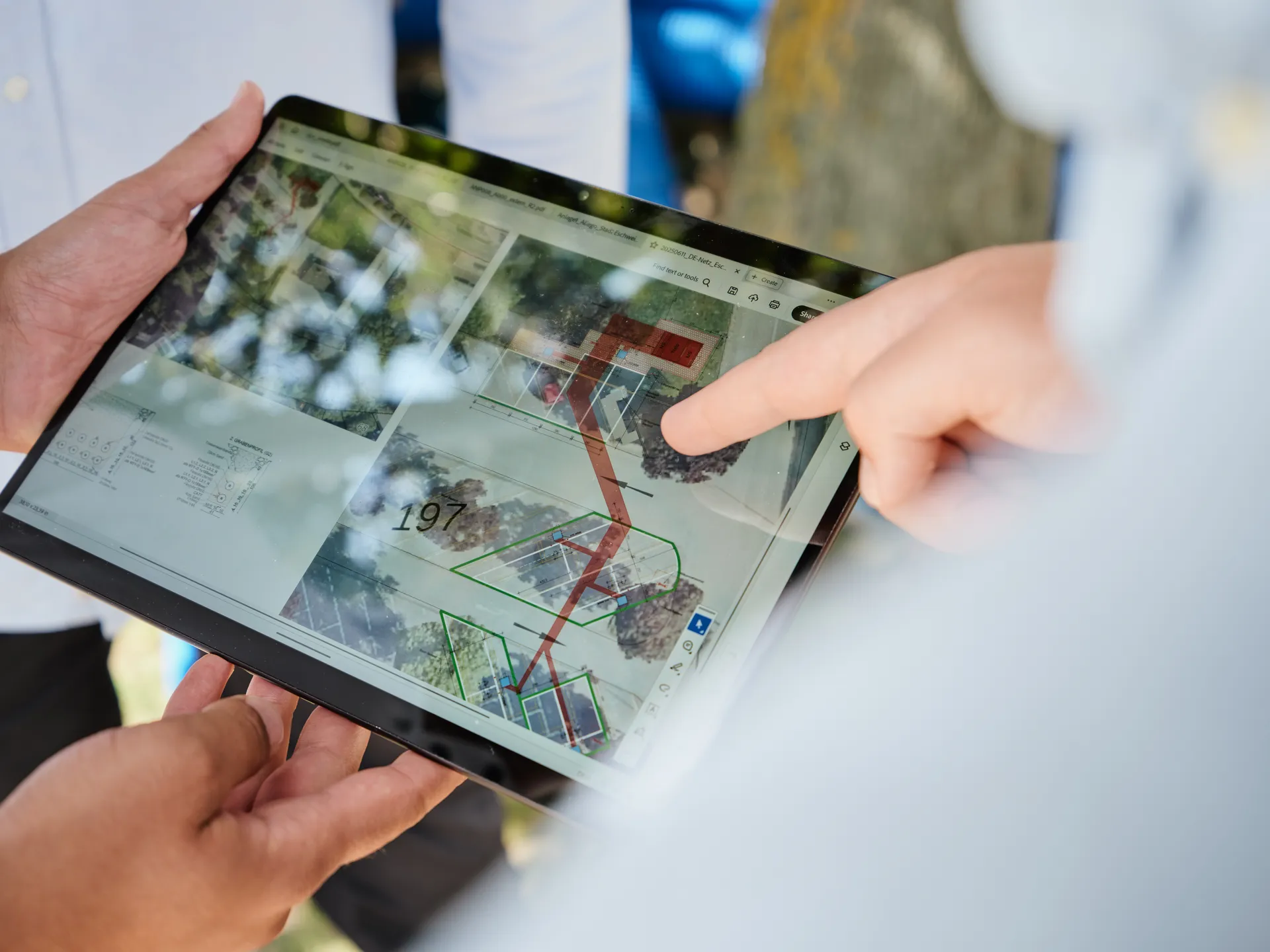 Nahaufnahme eines Tablets, das von zwei Personen gehalten wird. Auf dem Bildschirm ist ein digitaler Ausführungsplan mit Luftbildern und eingezeichneten Linien zu sehen. Eine Person zeigt mit dem Finger auf einen Punkt des Plans.