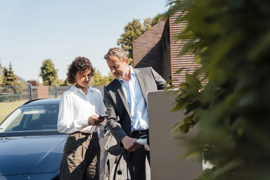 Eine Frau mit lockigen Haaren und weißer Bluse steht neben einem Mann mit grauem Sakko. Sie hält ein Smartphone, er das Ladekabel einer Ladesäule. Im Hintergrund steht ein blaues E-Auto, dahinter sind moderne Holzgebäude und Bäume zu sehen.