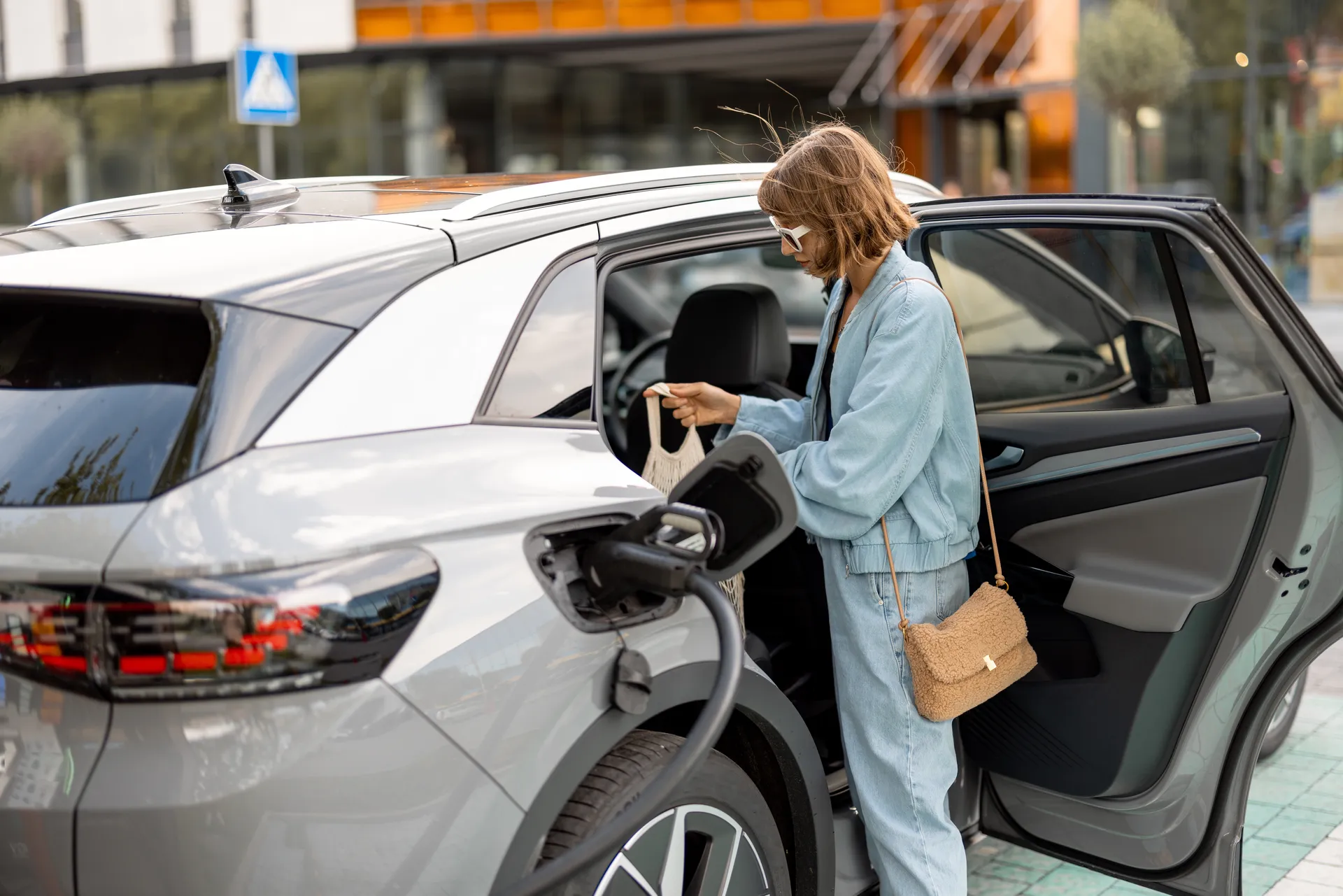 Eine Frau steht neben einem grauen Elektroauto mit geöffneter hinterer Tür und angeschlossenem Ladekabel. Sie packt einen Einkaufsbeutel ins Auto; im Hintergrund sind ein Gebäude und ein Fußgängerüberweg-Schild zu sehen.