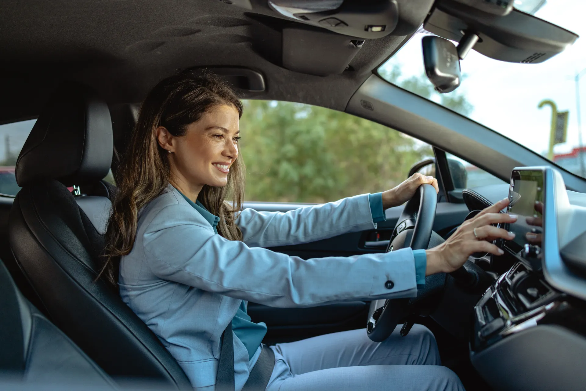 Eine junge, lächelnde Frau mit langen braunen Haaren sitzt im Fahrersitz eines modernen Autos, trägt einen hellblauen Blazer und bedient mit der rechten Hand den Touchscreen des Infotainmentsystems.