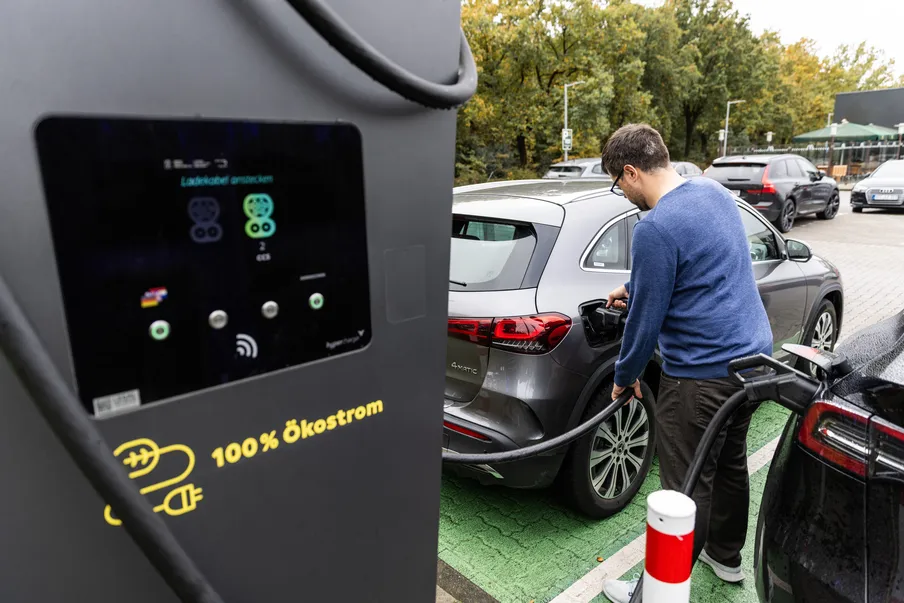 Ein Mann schließt ein graues Elektroauto an eine schwarze Ladesäule an, die "100% Ökostrom" anzeigt.